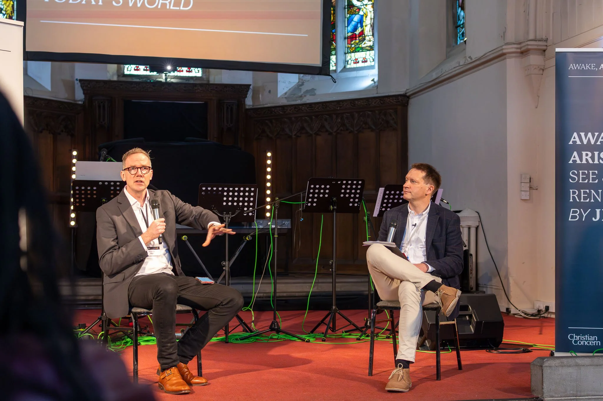 Two men are sitting and talking in a church with stained glass windows. One is speaking into a microphone, wearing glasses and a suit. The other is sitting cross-legged, holding a microphone, wearing beige pants and a blazer. There are music stands, 