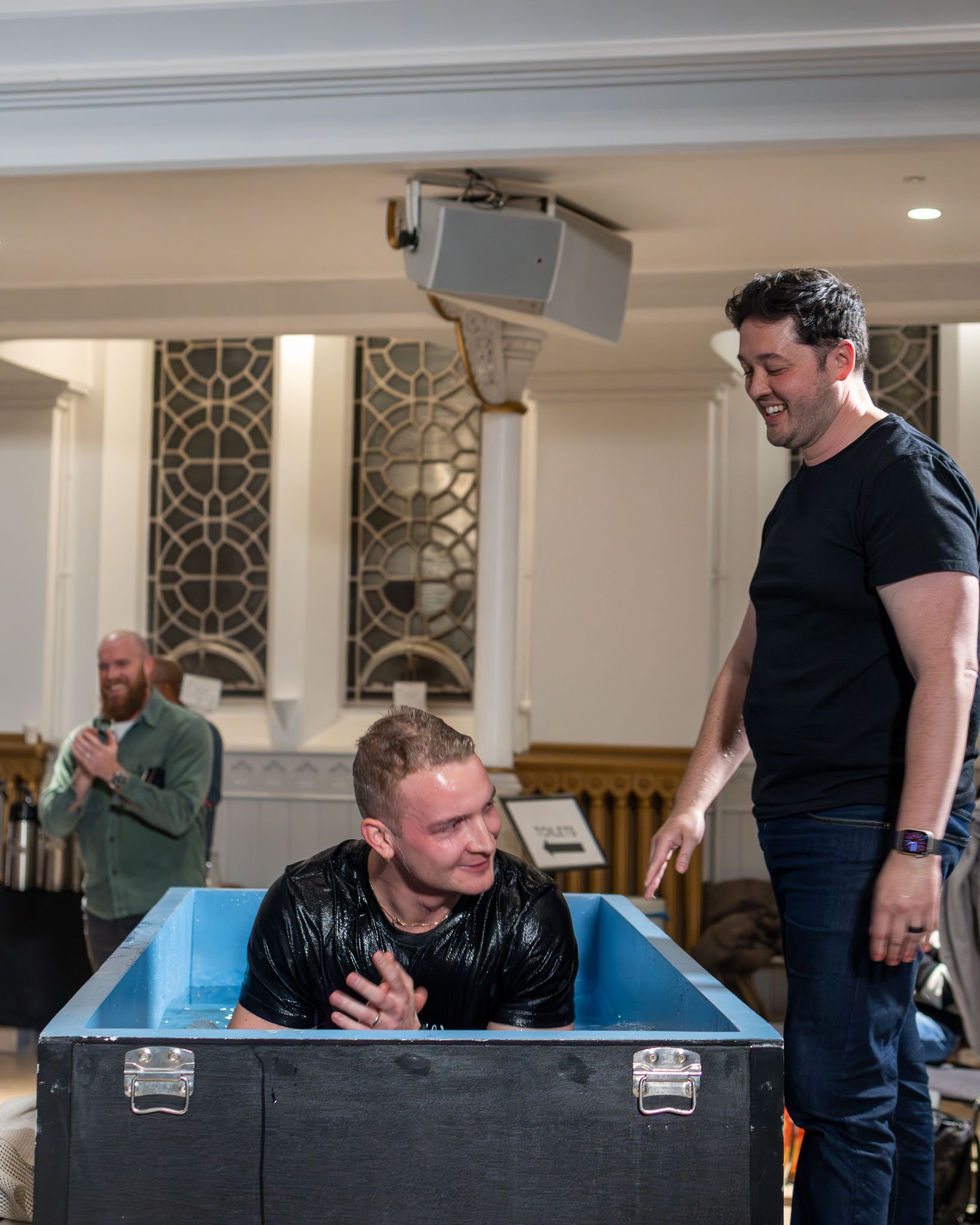 A man is being baptized in a large blue water container at an indoor church service, with another man standing nearby smiling.