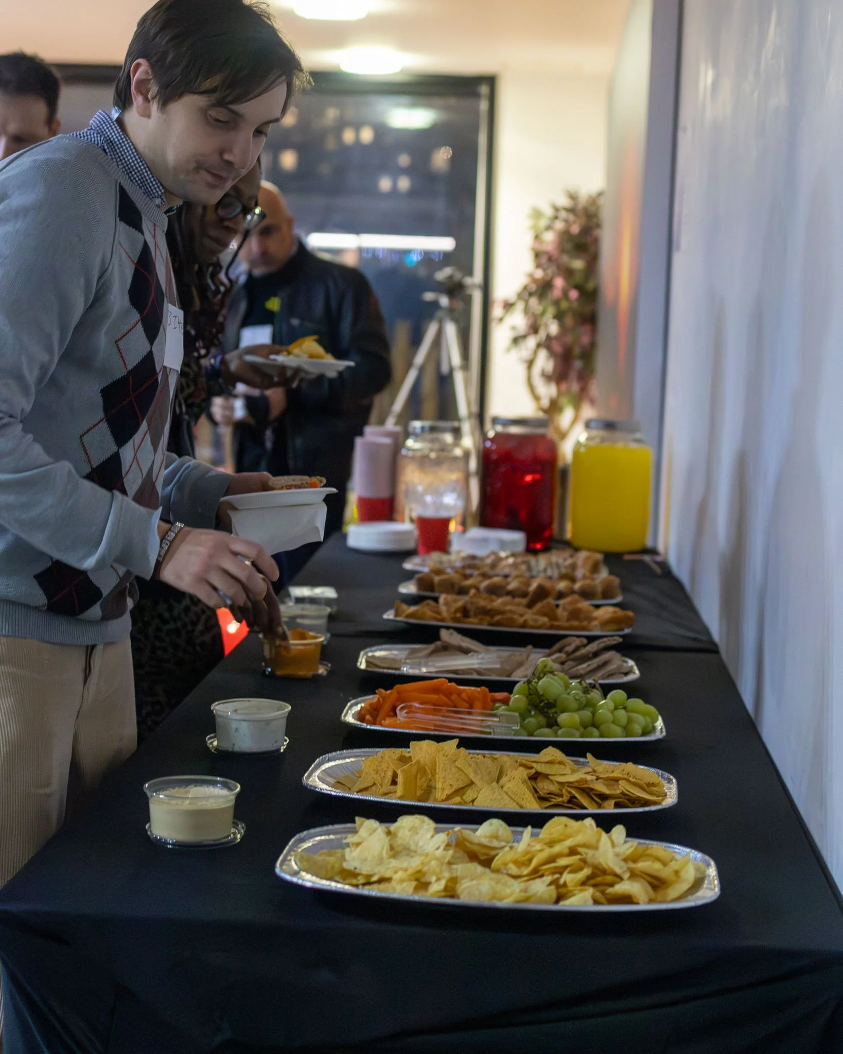 Buffet table with various snacks such as chips, vegetables, grapes, and dips, in a well-lit indoor setting with several people serving themselves.