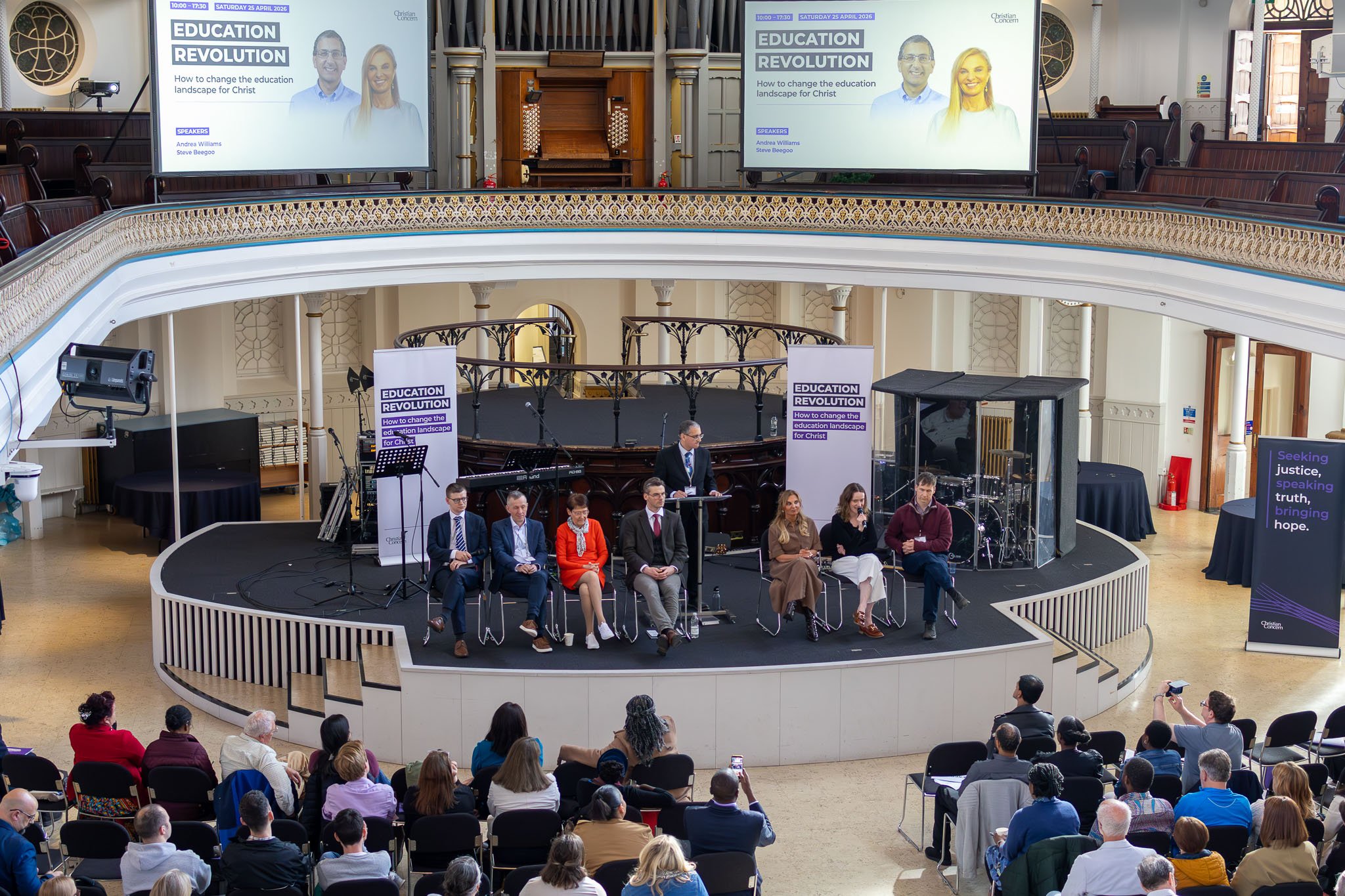 A conference or panel discussion titled 'Education Revolution' with speakers Andrea Williams and Steve Beegoo, held in a historic building with a balcony, screens, and a stage, attended by an audience.