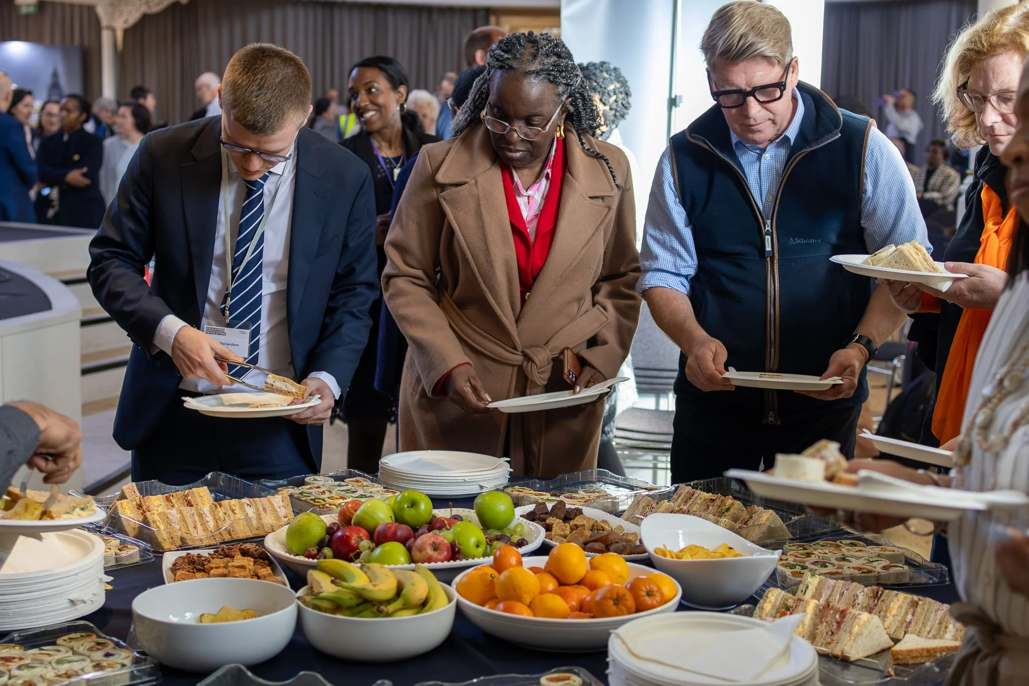 People serving themselves food at a buffet table with various dishes, including sandwiches, fruits, and desserts, at a social event or conference.