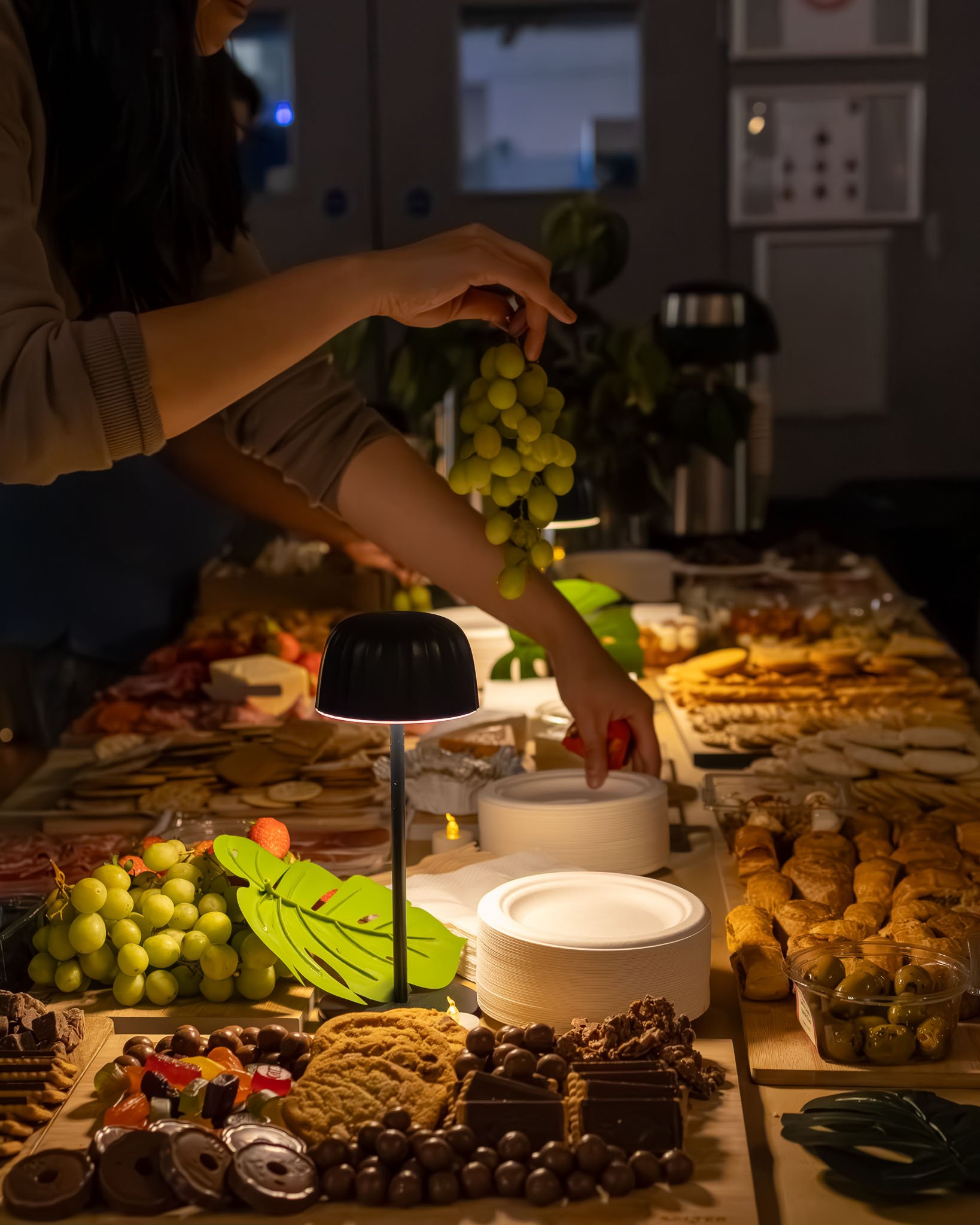 A dessert table filled with various sweets, including chocolates, cookies, and fruit, with a person reaching for food. There are plates, bowls, and food items on the table, illuminated by a nearby lamp, with a dimly lit room in the background.