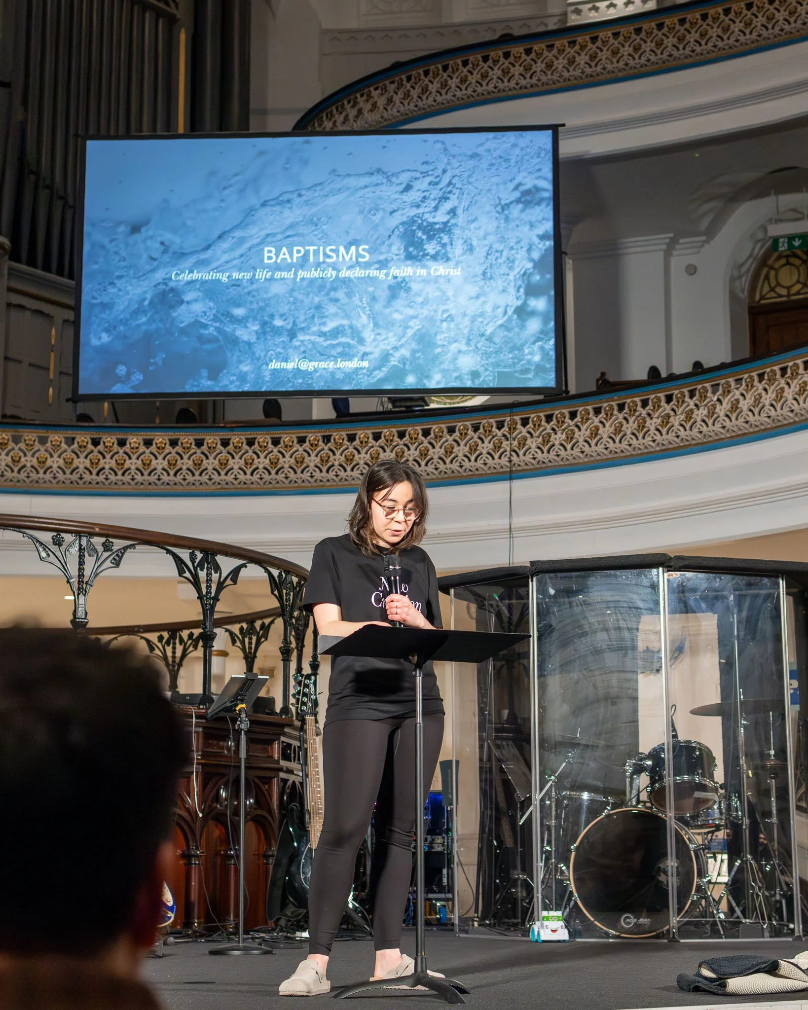 A woman speaking at a podium during a baptism event in a church, with a large video screen behind her displaying the word 'BAPTISMS' and the phrase 'Celebrating new life and publicly declaring faith in Christ' along with an email address.