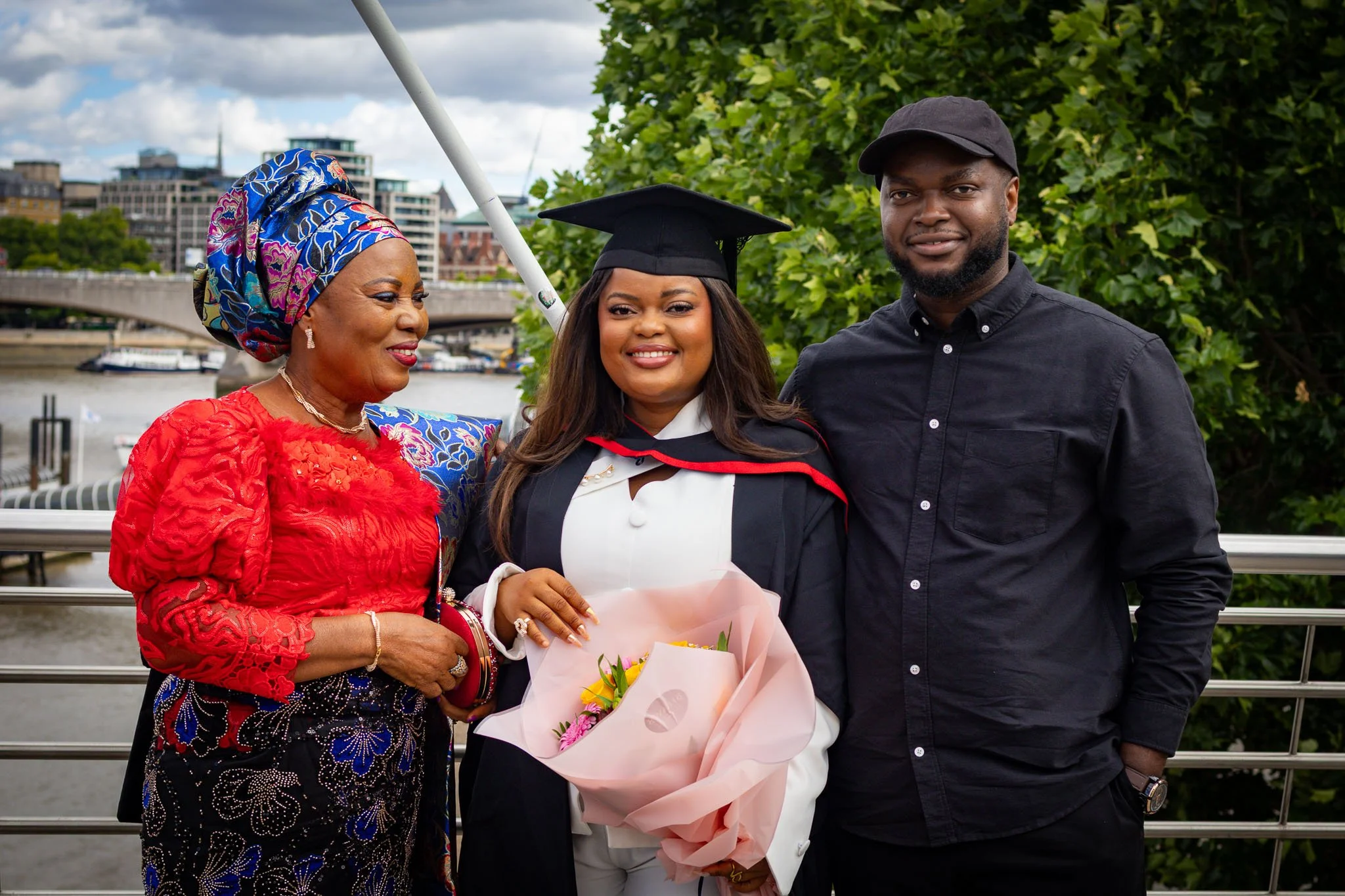 A woman in graduation cap and gown smiling with a woman in red and purple patterned blouse and a man in black shirt and cap, standing outdoors near a river and city buildings, with greenery in the background, during daytime.