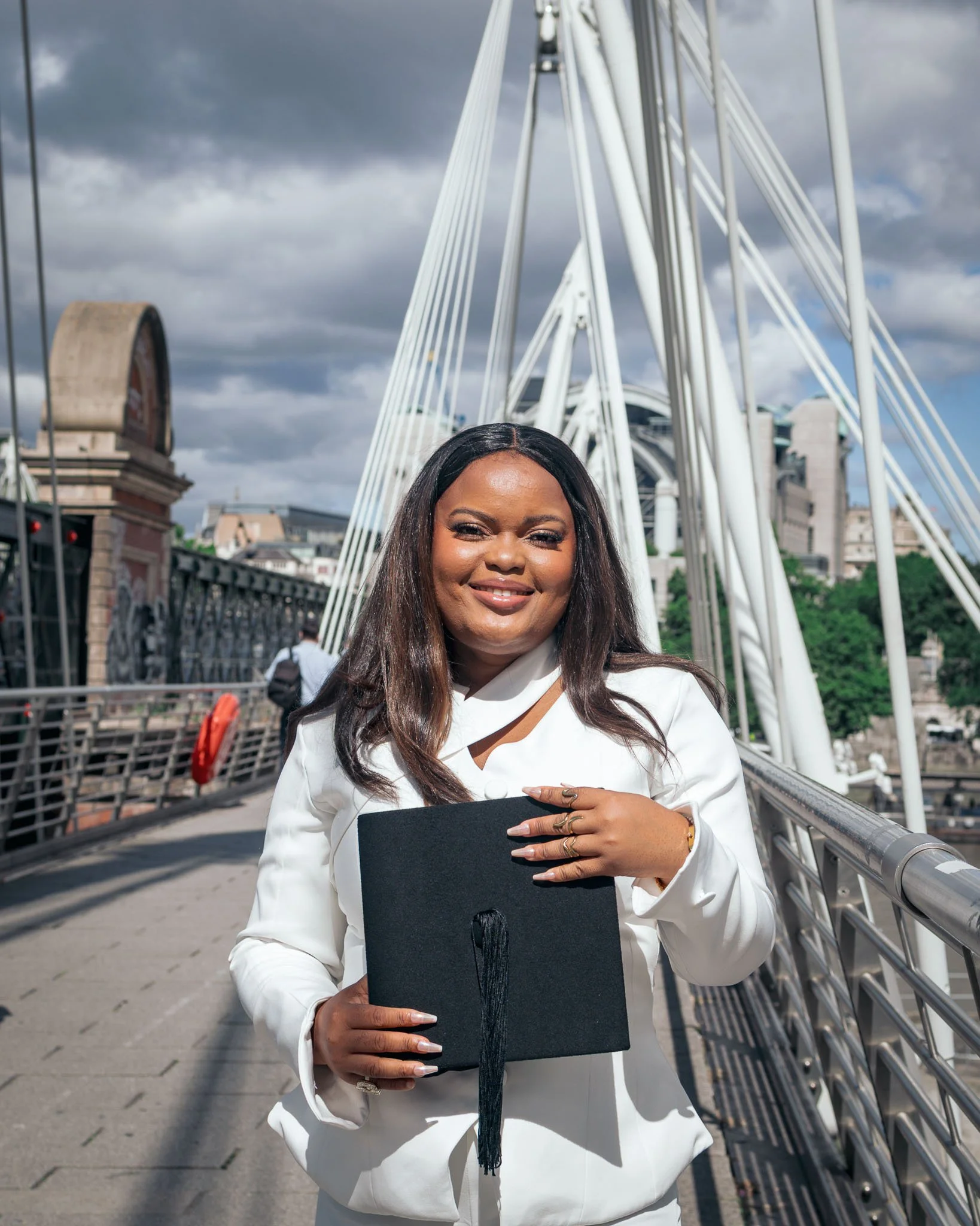 A woman in a white suit holding a black graduation cap with a tassel, standing on a bridge with a cityscape and cloudy sky in the background.