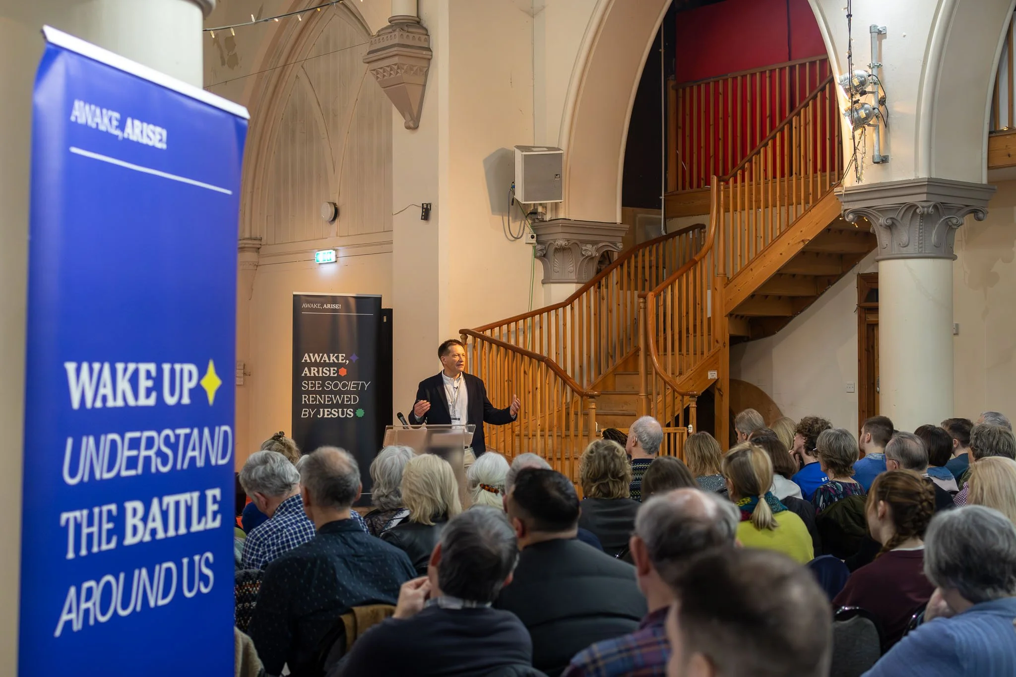 A man in a suit giving a speech at a podium during a conference or seminar, with an audience seated facing him. The room has wood accents, a staircase, and banners with text including "Awake, Arise!" and "See Society Renewed by Jesus." A large blue b