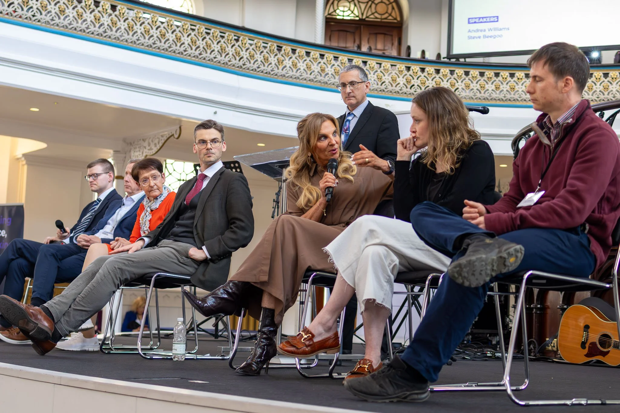 A group of diverse people sitting on stage in a conference or panel discussion at an indoor venue, with a woman speaking into a microphone and others listening attentively.