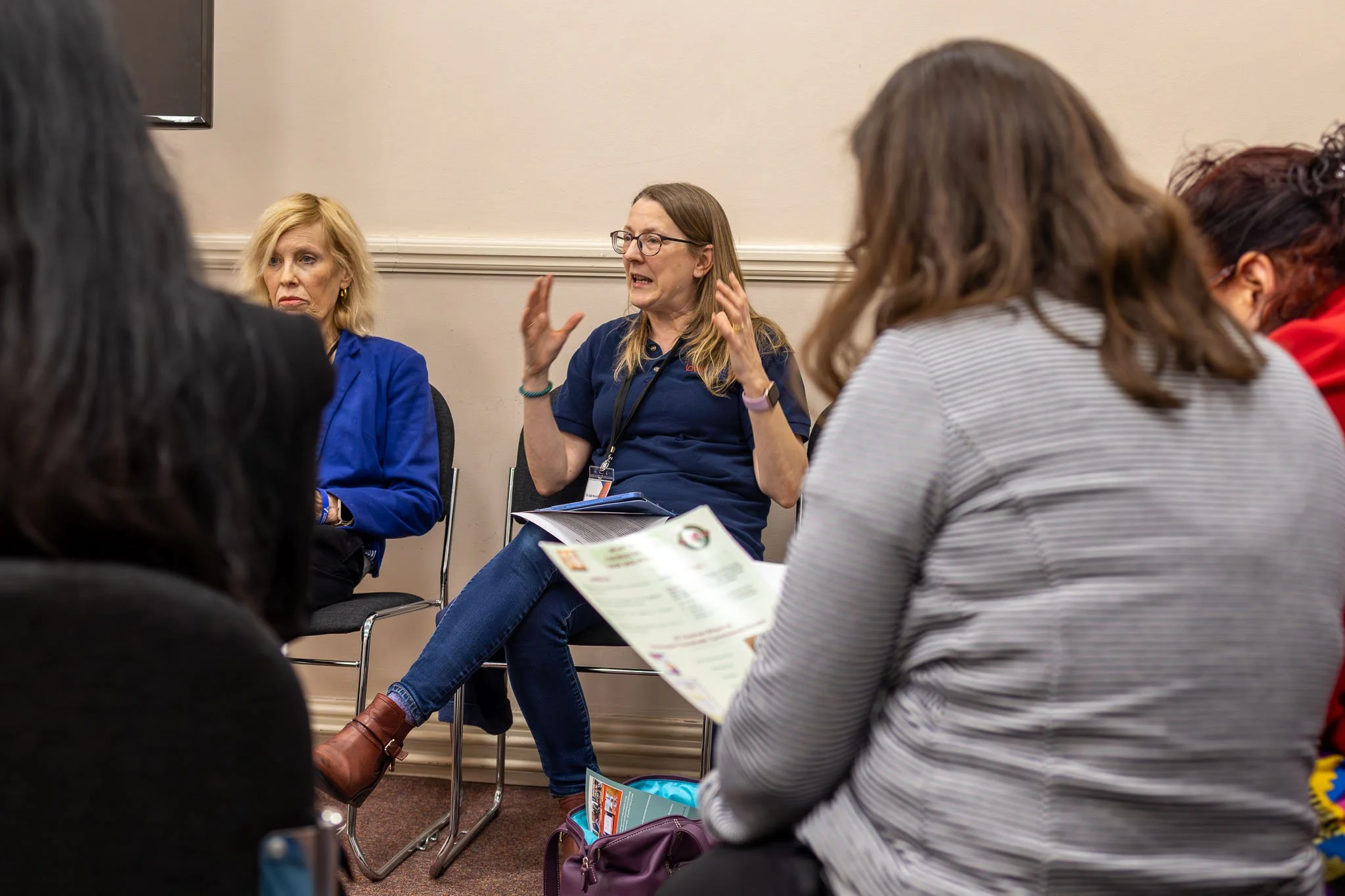 A woman with glasses and long hair is speaking passionately during a discussion in a small group setting. She is gesturing with her hands while sitting on a chair, with a notebook on her lap. Several other people are sitting around her, some holding 