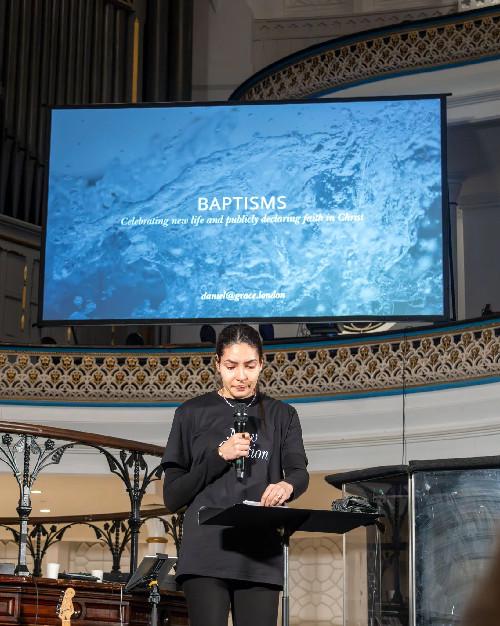 A woman speaking into a microphone during a baptism celebration, with a large screen above displaying the word 'BAPTISMS' and the phrase 'Celebrating new life and publicly declaring faith in Christ'.