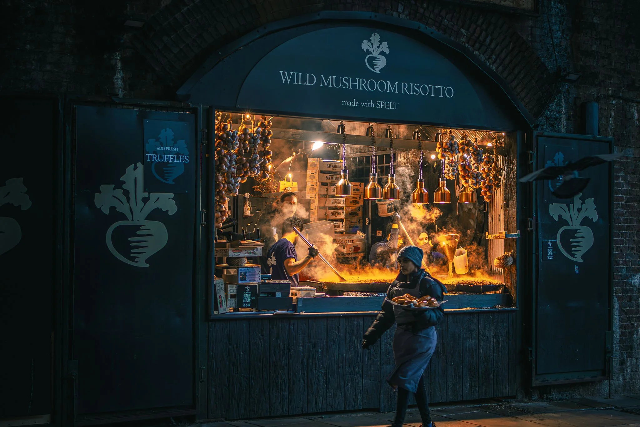 At Borough Market, workers are selling wild mushroom risotto made with spelt, with decorative garlic and herbs hanging above the counter. A worker is preparing food inside, and a customer is taking a plate of food outside.