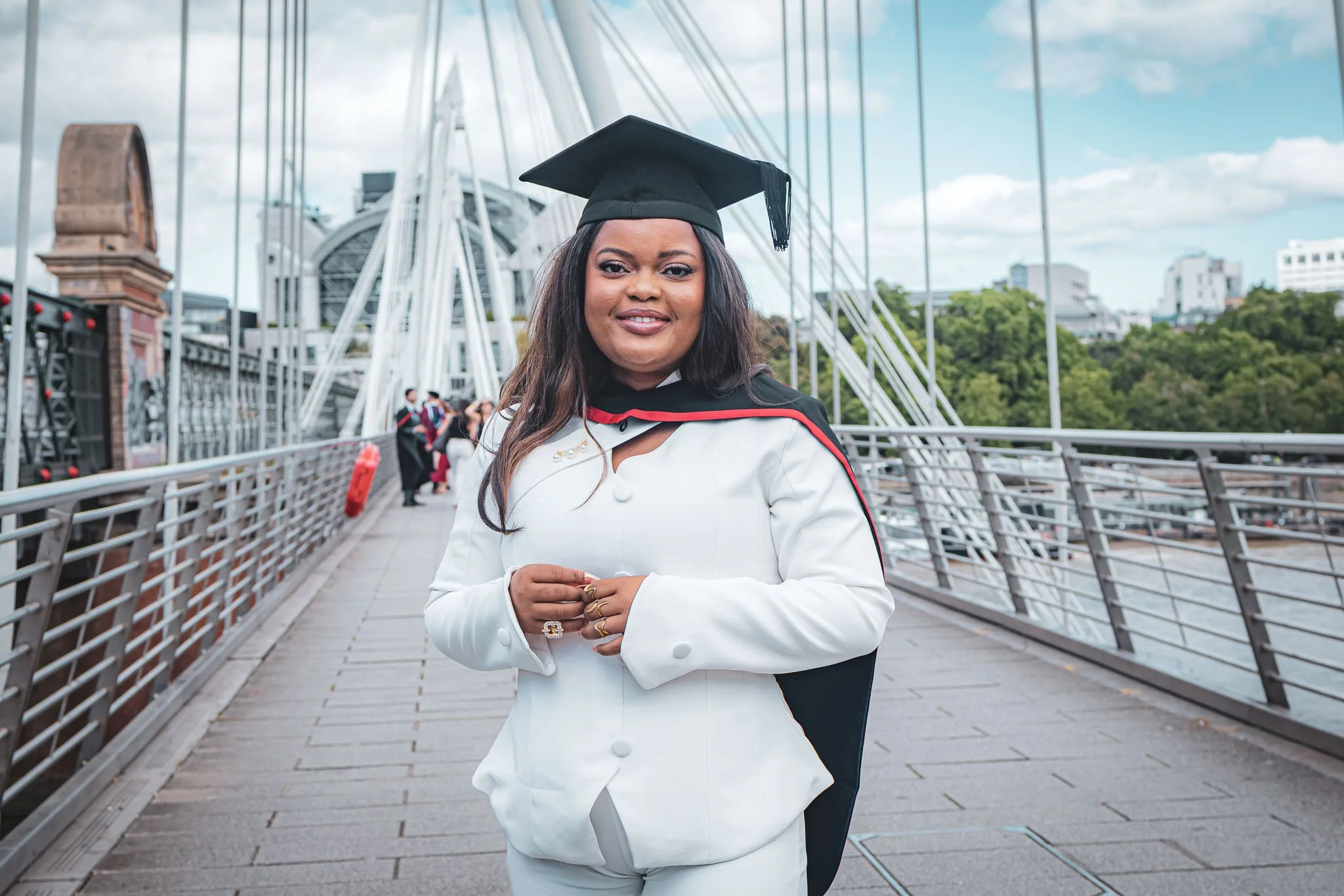 Woman in white suit and graduation cap standing on a bridge with a cityscape background.