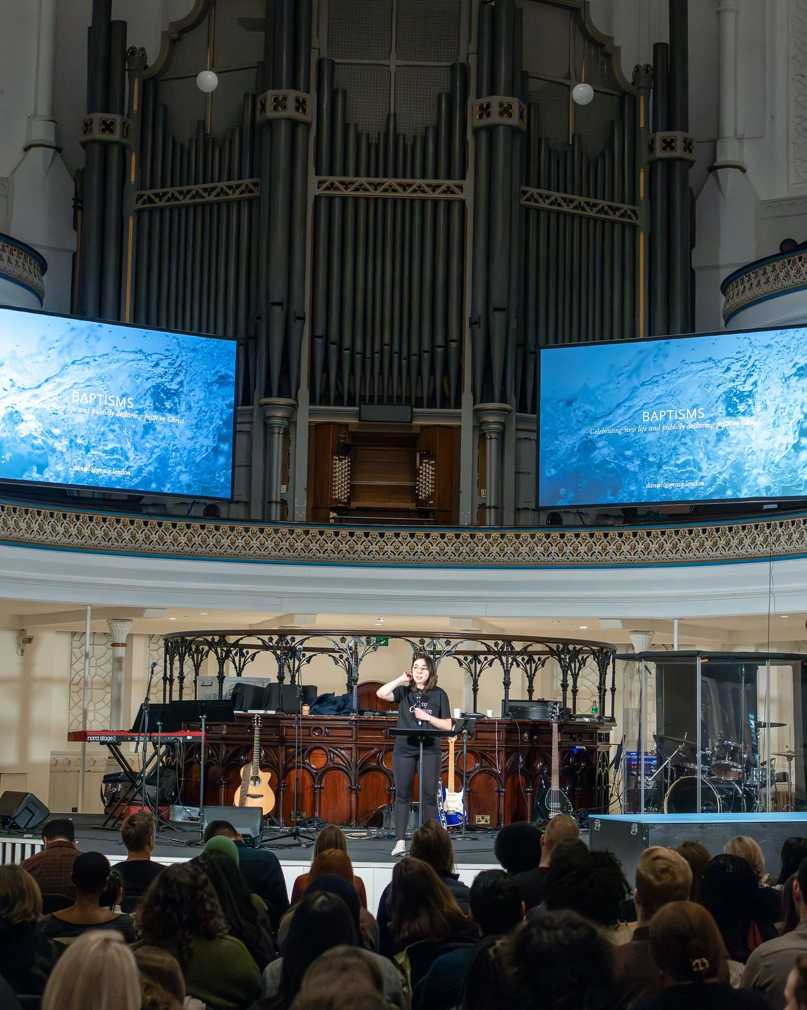 A woman standing on stage speaking to an audience in a church or concert hall, with musical instruments and microphones around her, and a large pipe organ behind her. The audience is seated and listening attentively.