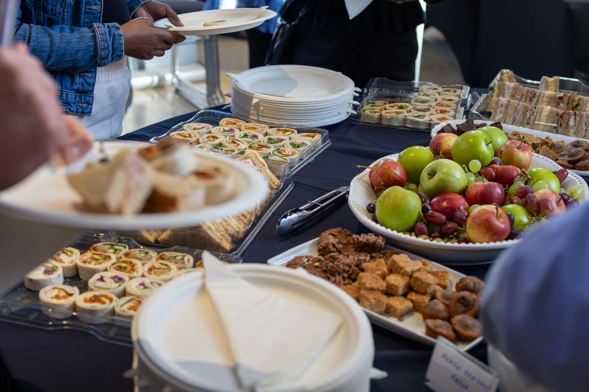 People selecting food at a buffet table with trays of mini sandwiches, fresh apples, brownies, and other baked goods.