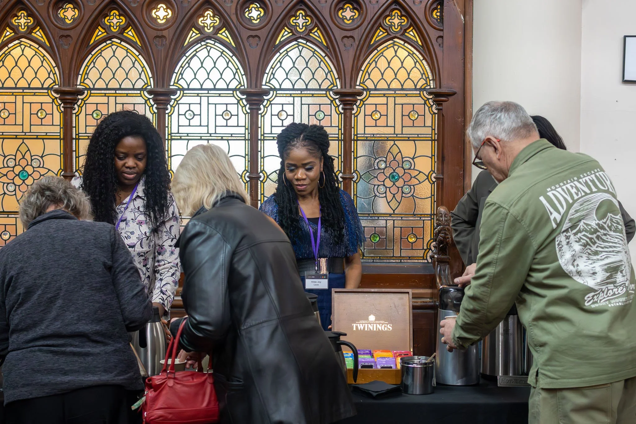 People standing at a counter with Twinings tea products, in front of a decorative stained glass window.