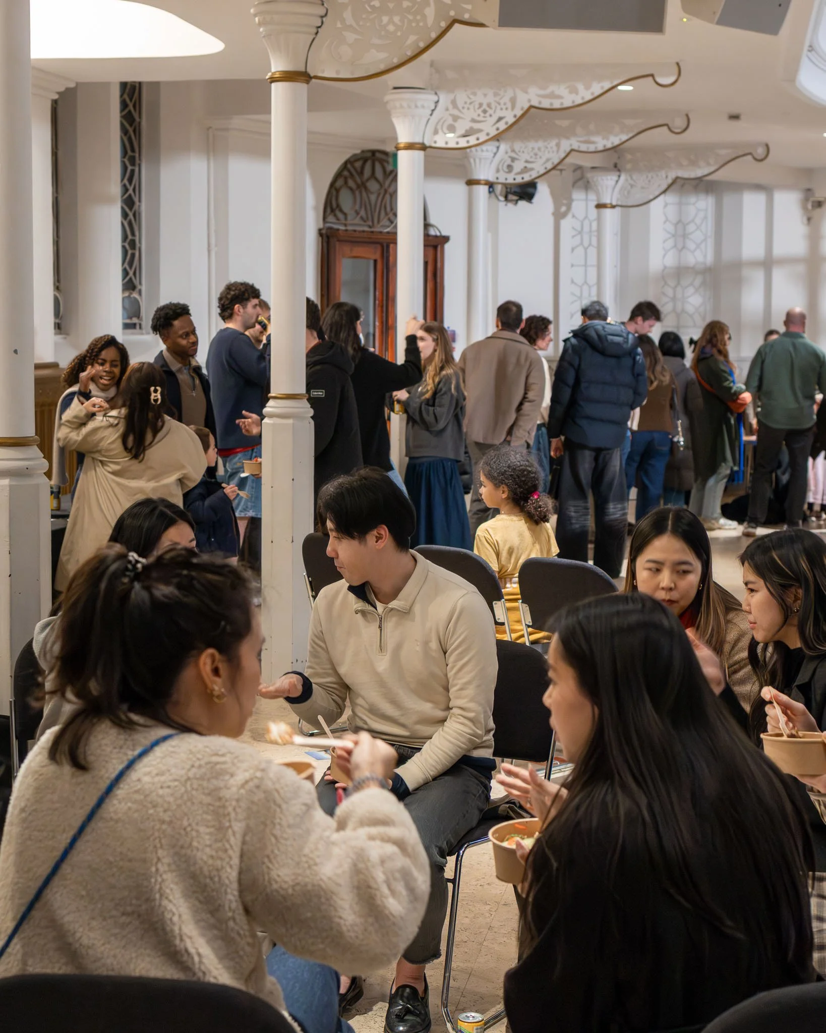 People gathered in a large indoor space, some are seated at tables eating, others stand in line or chat, with ornate white columns and decorative ceiling fixtures in the background.