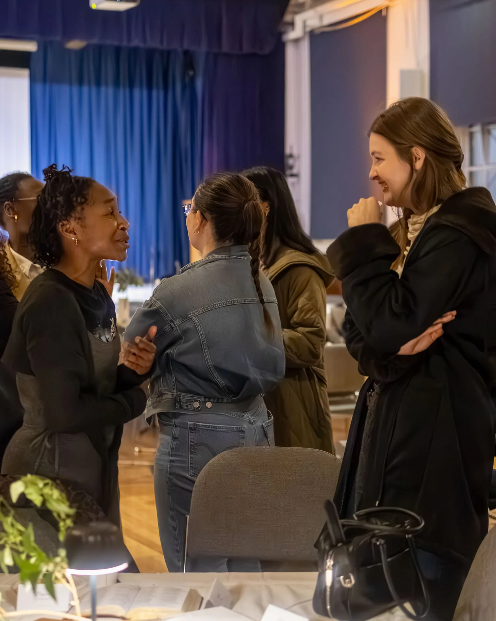 A group of women engaged in conversation in a room with blue curtains and wooden floors, with some women smiling and laughing.