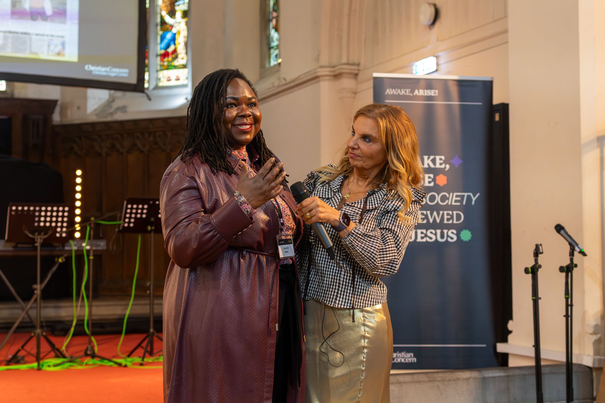 Two women conversing at an indoor event, one holding a microphone. Behind them, there is a sign that says "AWAKE, ARISE!" and stained glass windows.