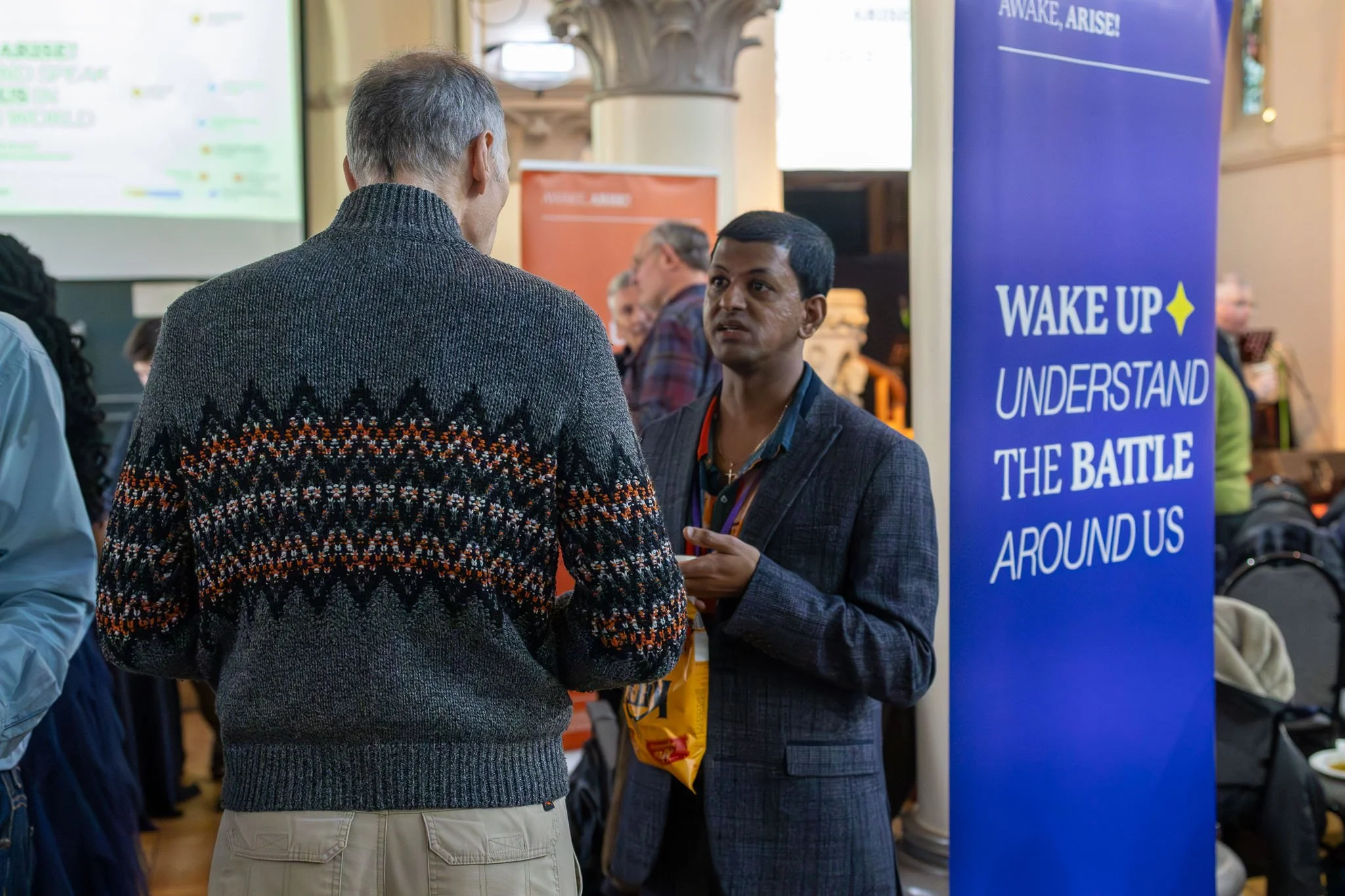 Two men engaged in conversation at an indoor event, with a large blue banner in the background that reads, 'Wake Up, Understand the Battle Around Us.' Several other people are visible in the background.