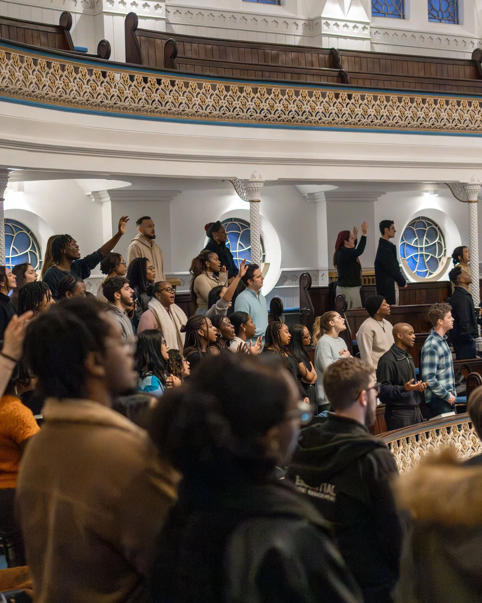 People gathered inside a church, listening attentively, some raising their hands in response to worship. The architecture includes ornate, decorative balcony railings and arched windows with stained glass.