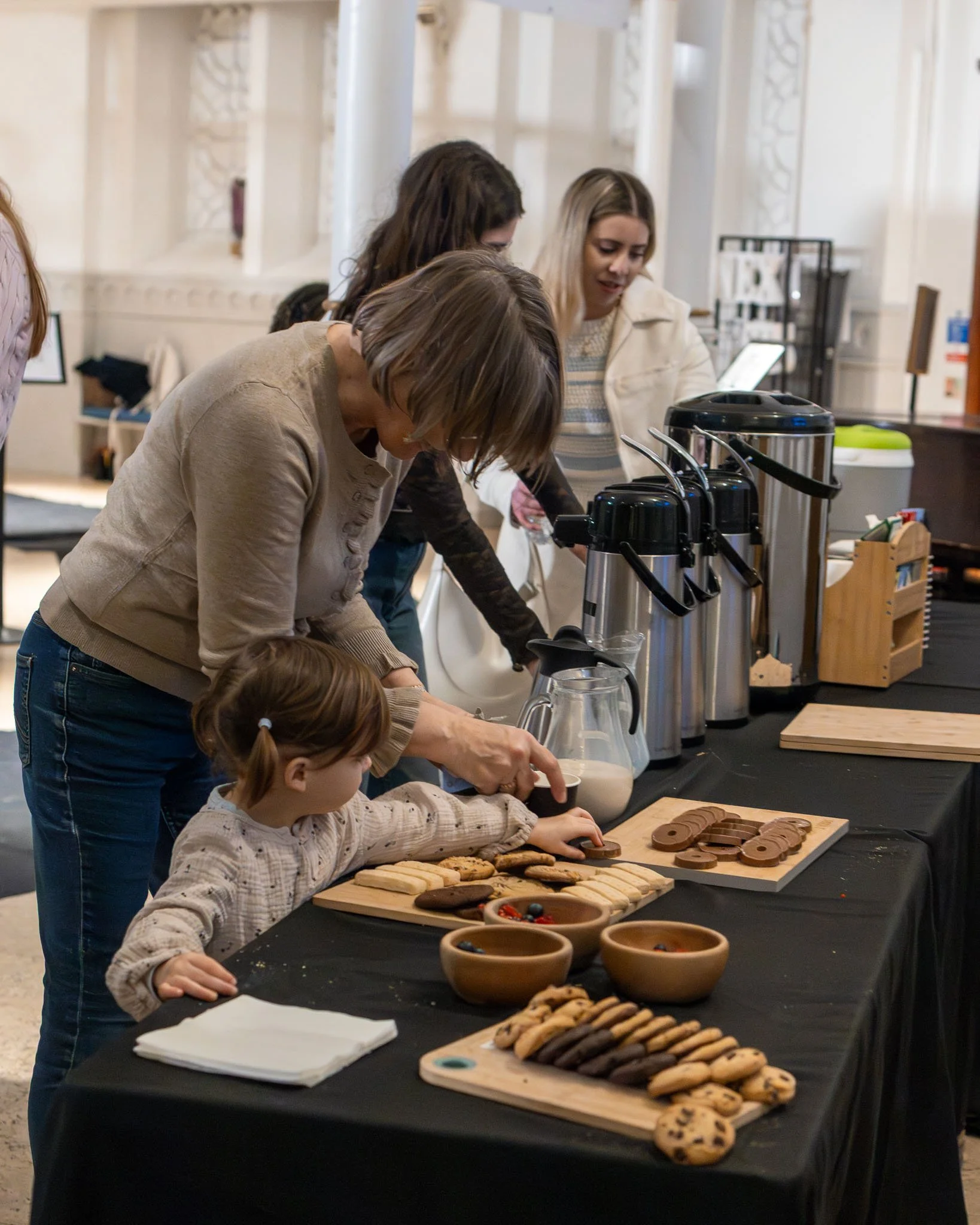 A group of people, including children and adults, are gathered around a table with baked cookies and bowls of toppings, participating in a cookie decorating activity in a bright, decorated room.
