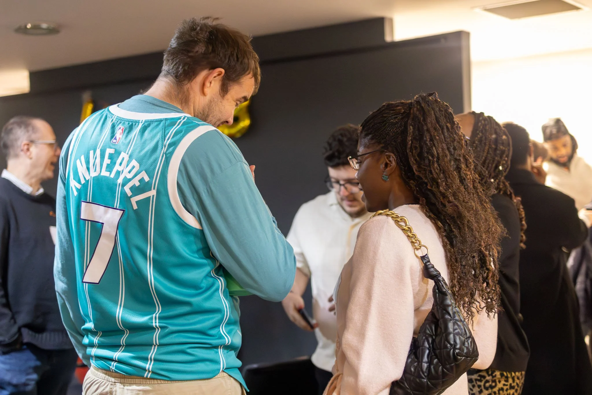 A man wearing a teal basketball jersey with the name 'Anders' and the number 7 is talking to a woman with long, curly brown hair and glasses at a social gathering. Other people are visible in the background.