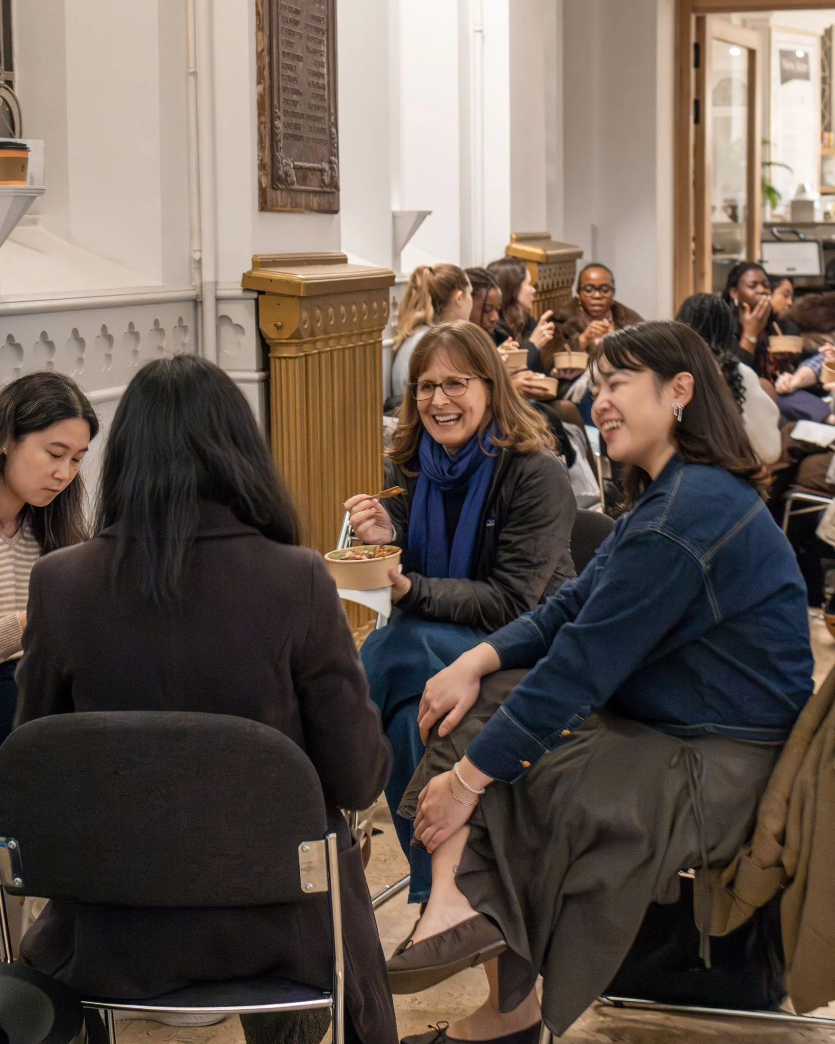 People sitting in a circle at a social gathering, engaging in conversation and holding bowls of food, in an indoor setting with warm lighting and wooden decor.