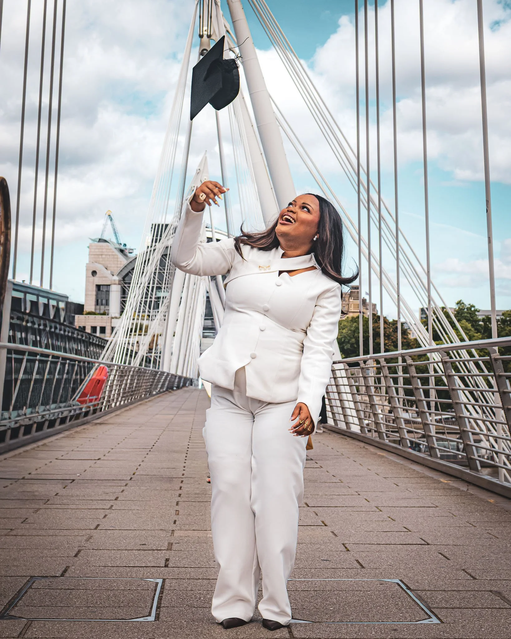 A woman in a white suit celebrating graduation on a bridge in London with a graduation cap thrown in the air.