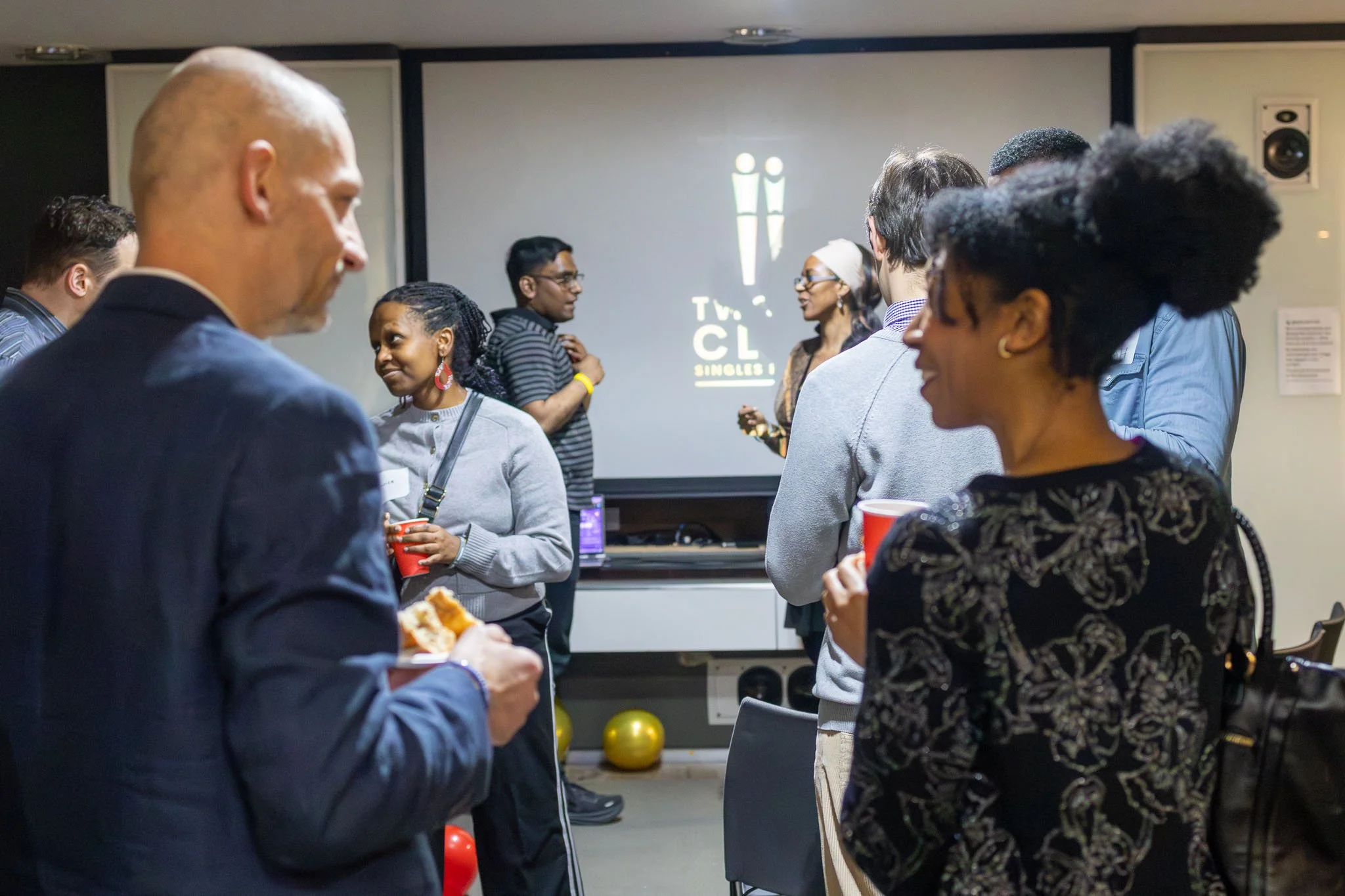 People gathered in a room at a social event, engaging in conversations, with a TV channel logo projected on the wall in the background.