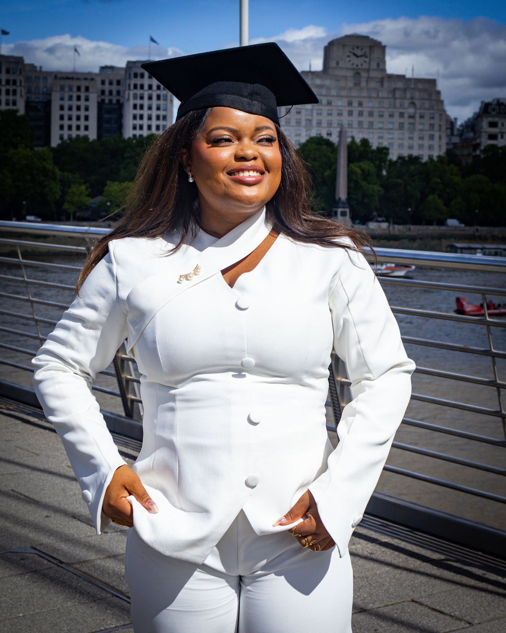 Woman in white suit and black graduation cap smiling outdoors near waterfront with city buildings and cloudy sky in background.