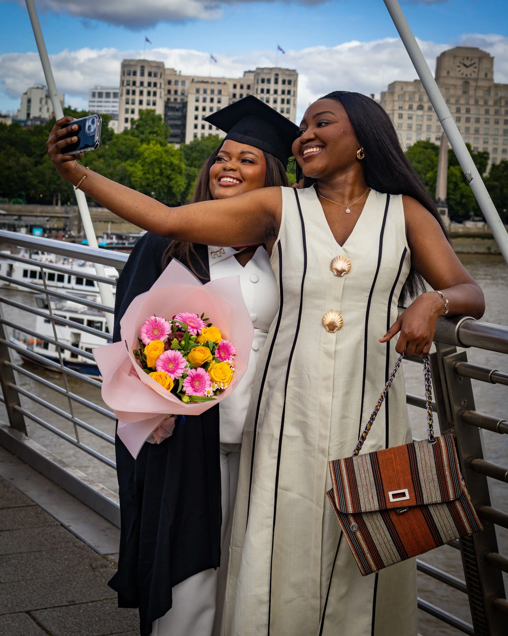Two women taking a selfie on a bridge in London. One woman is wearing a graduation cap and gown, and the other woman is dressed in a beige dress. They are smiling and holding a bouquet of flowers. 