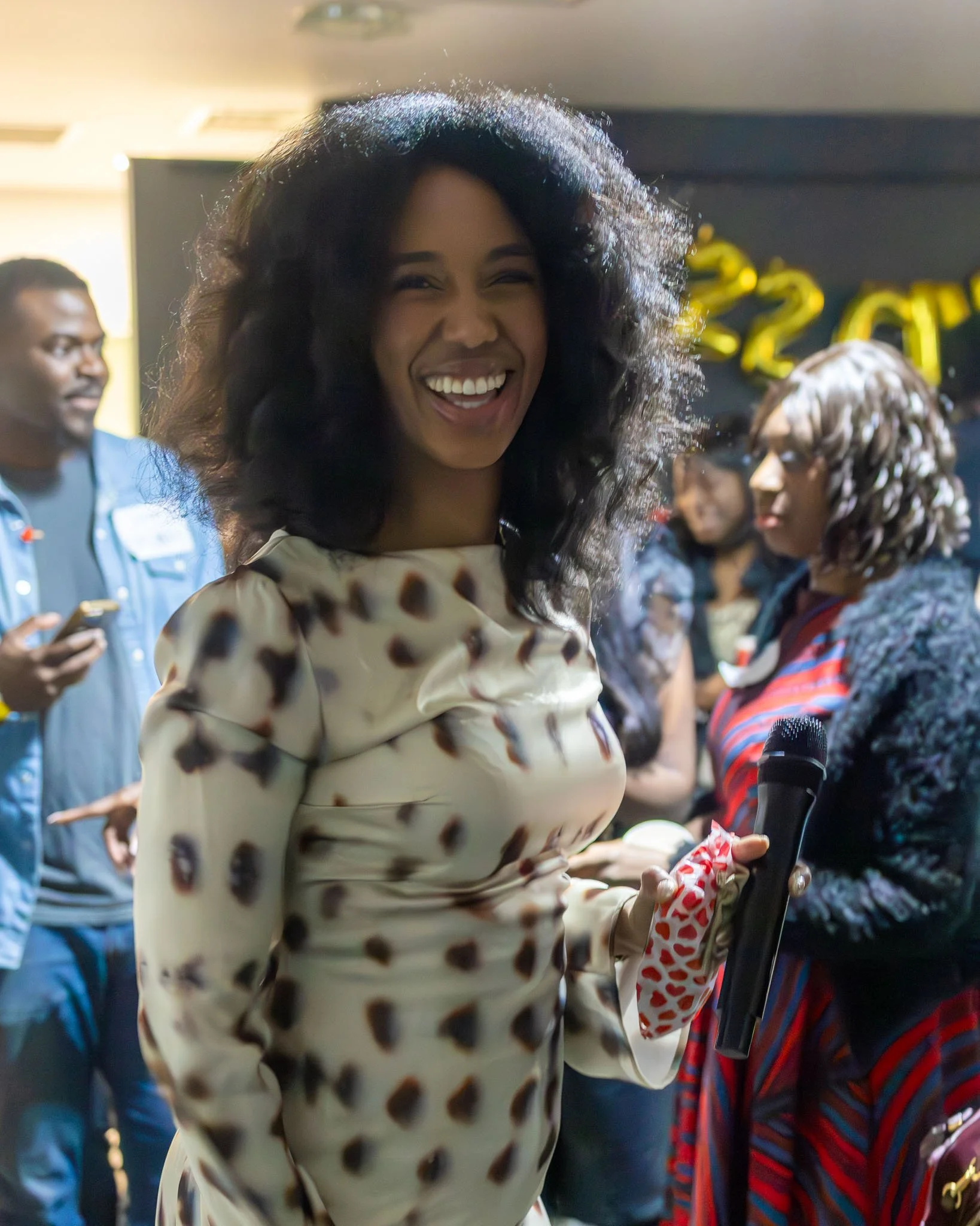 A woman with curly hair smiling and looking into the camera, wearing a polka dot dress at a celebration, with balloons and people in the background.