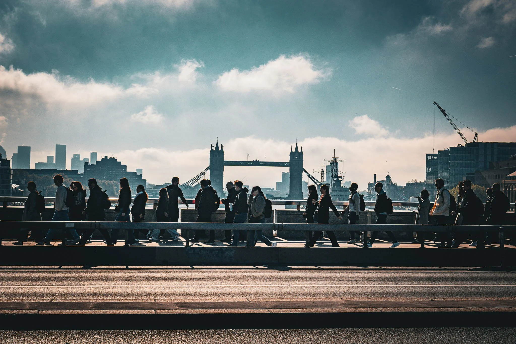 People walking across a bridge with the London Tower Bridge in the background, under a partly cloudy sky.