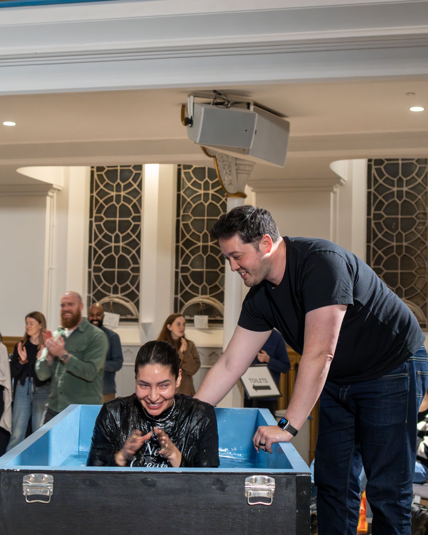 A woman with water on her face sitting in a blue tub, smiling, while a man stands beside her with a smile and a smartwatch, helping her wash her hair during a baptism ceremony with several onlookers in the background inside a decorated room.