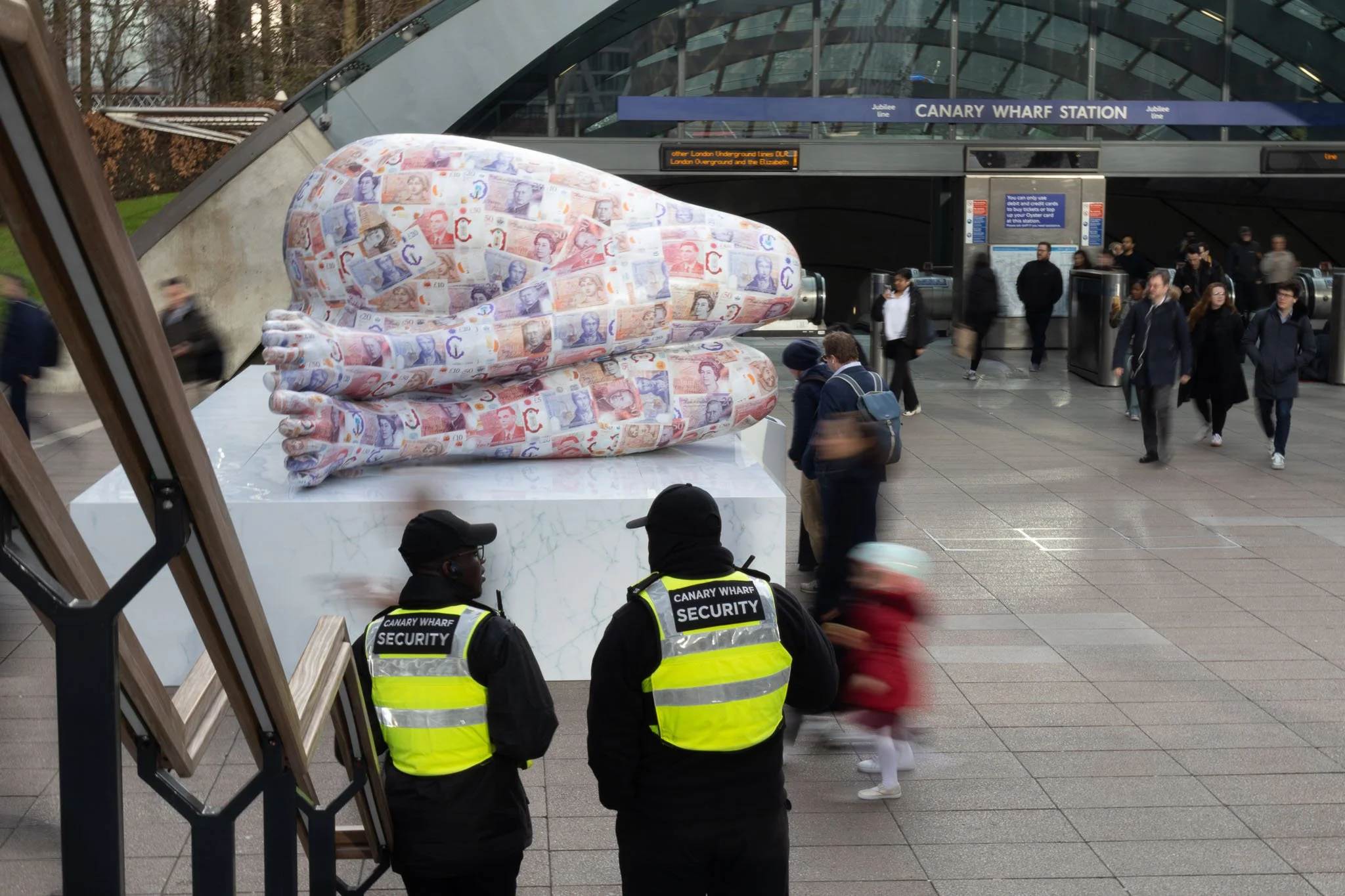 A large sculpture of a pair of lips covered in British pound banknotes, situated outside Canary Wharf Station. Several people walk past, and security personnel are visible in the foreground.