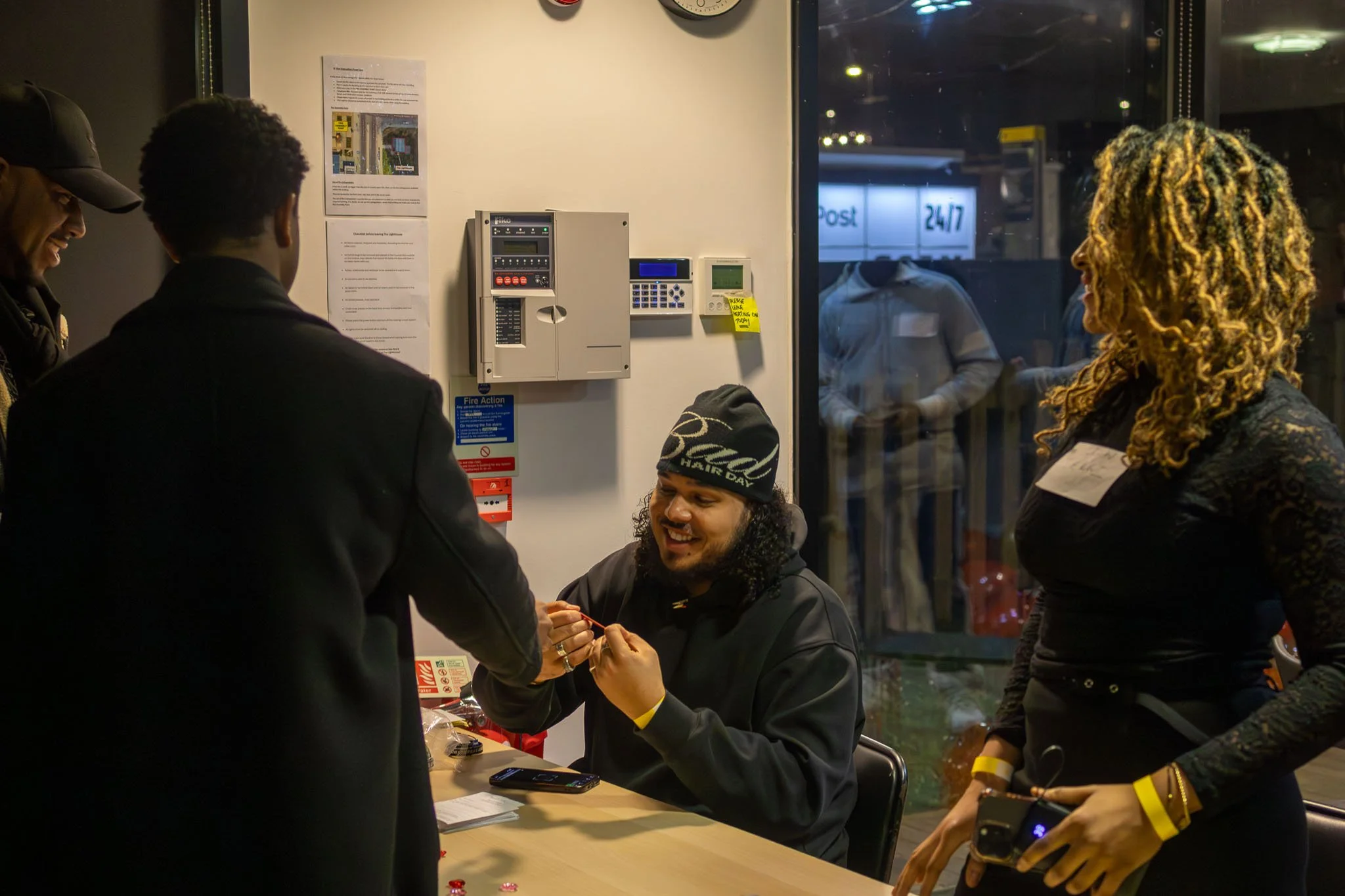 People gathered around a table inside a building at night, exchanging wristbands at an event,. There is a window showing streetlights and a sign that reads '24/7' outside.