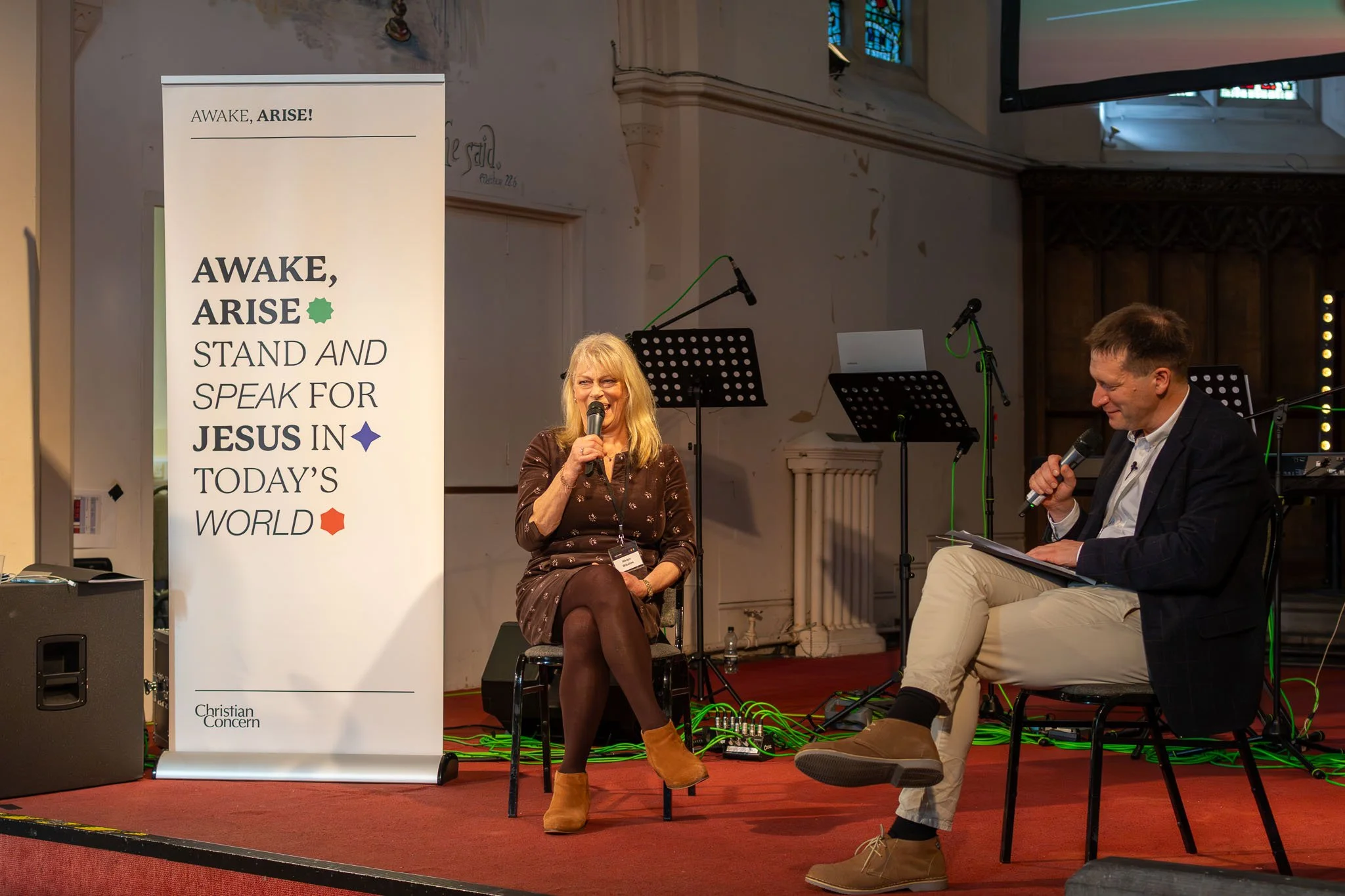 A woman with blonde hair sitting on a chair, speaking into a microphone, and a man with brown hair sitting next to her, holding a microphone and smiling. They are on a stage with musical equipment and a large banner that reads, 'Awake, Arise! Stand a