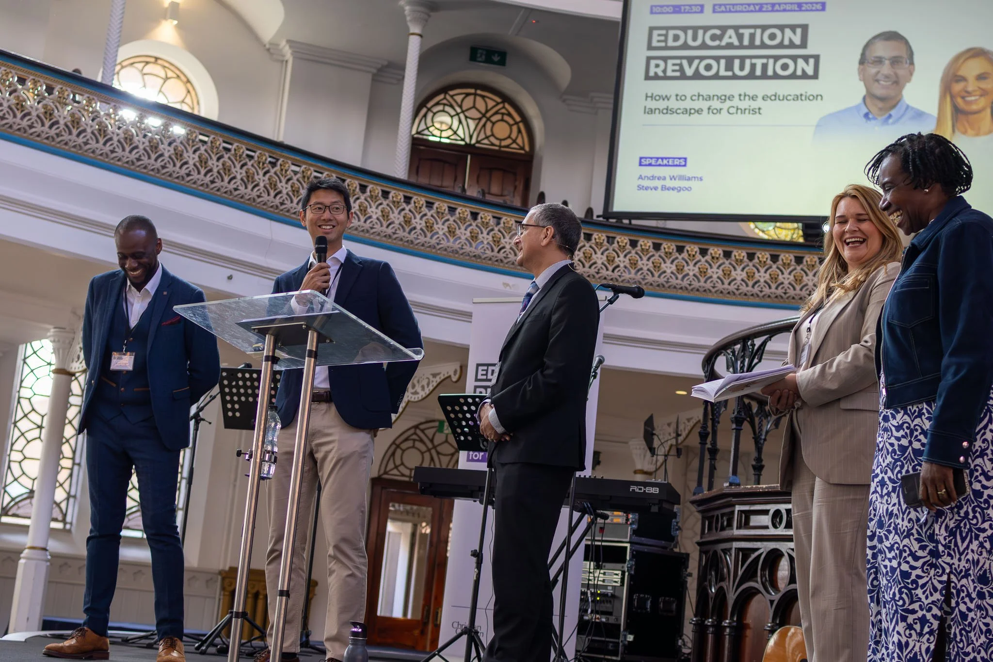 A group of diverse people standing and talking on stage at a conference titled 'Education Revolution' inside a building with ornate windows and railing, with a large screen displaying the conference title and speakers.