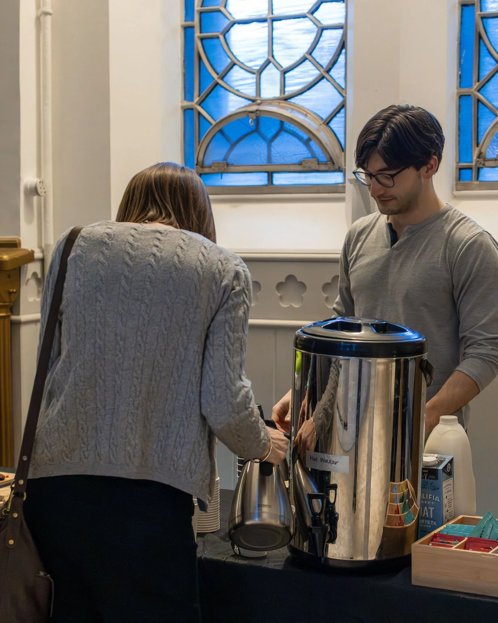 Two people at a table with a large metal container labeled 'Hot Water' and other supplies and products, inside a room with white walls and blue stained glass windows.