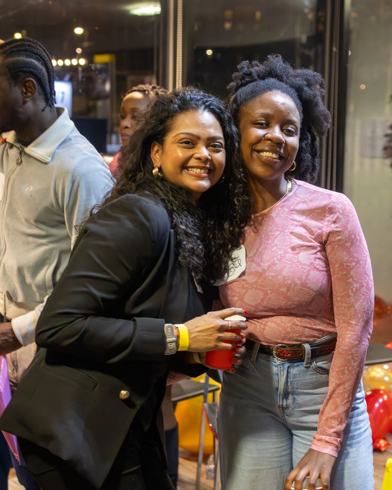 Two smiling women standing close together, one wearing a pink patterned top and jeans, holding a red cup, and the other wearing a black blazer with gold earrings and a watch, holding a brown beverage container. They are inside a room with glass walls