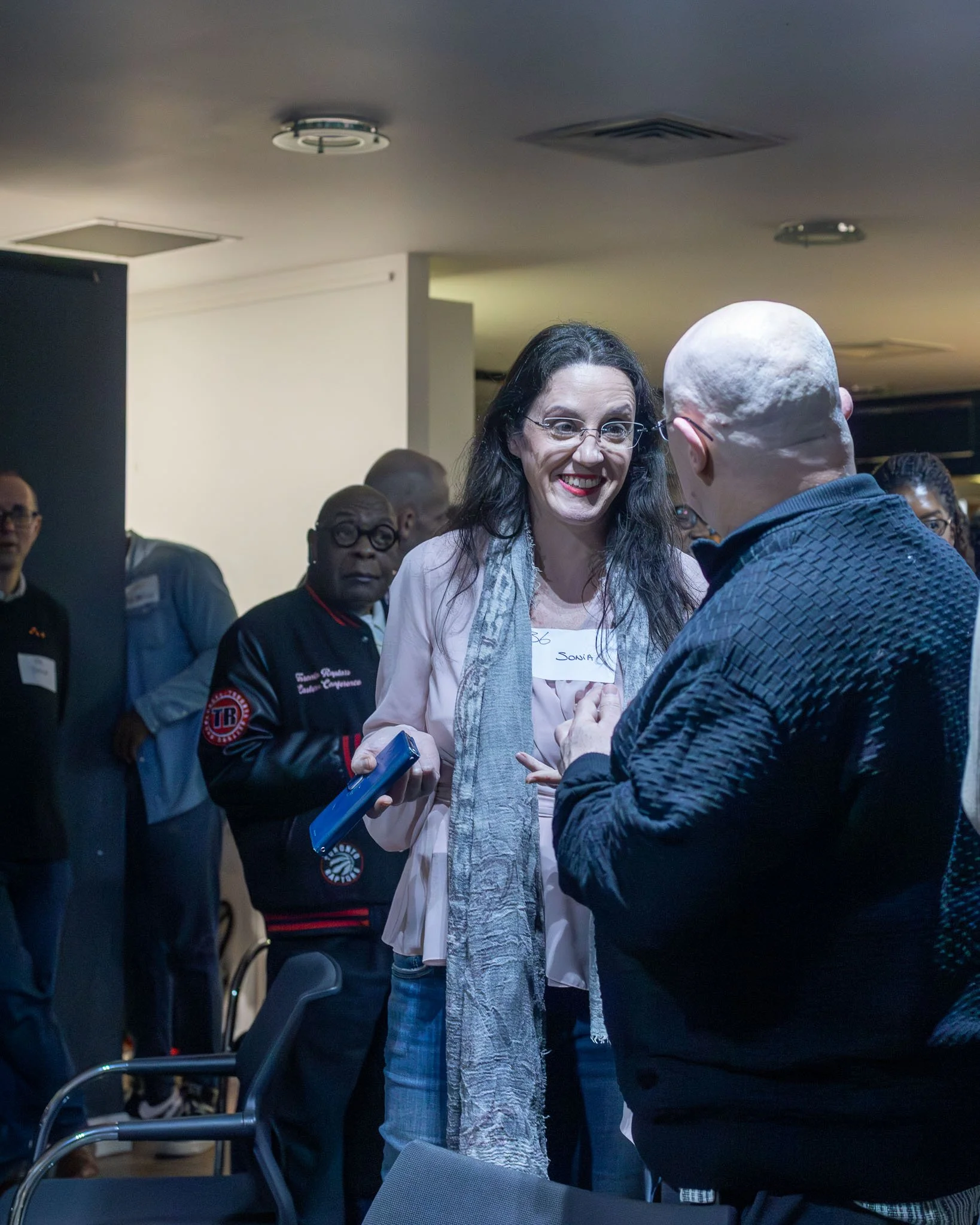 A woman with long dark hair, wearing glasses, a pink top, and a gray scarf, smiling and talking to a bald man in a dark jacket, with several people in the background at an indoor event.
