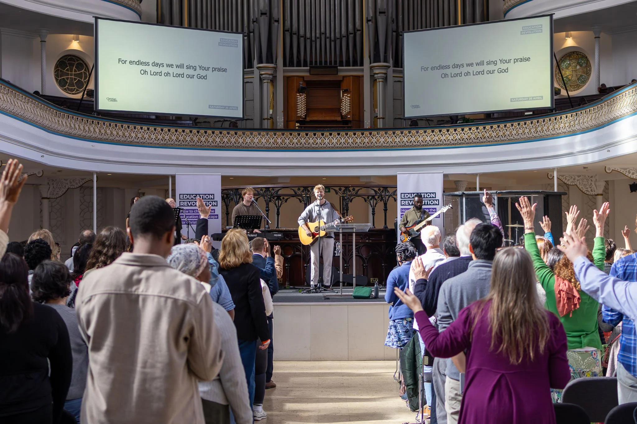 A church congregation gathered in a spacious sanctuary, raising their hands during a worship service with a band performing on stage, large screens displaying lyrics, and intricate architectural details in the background.