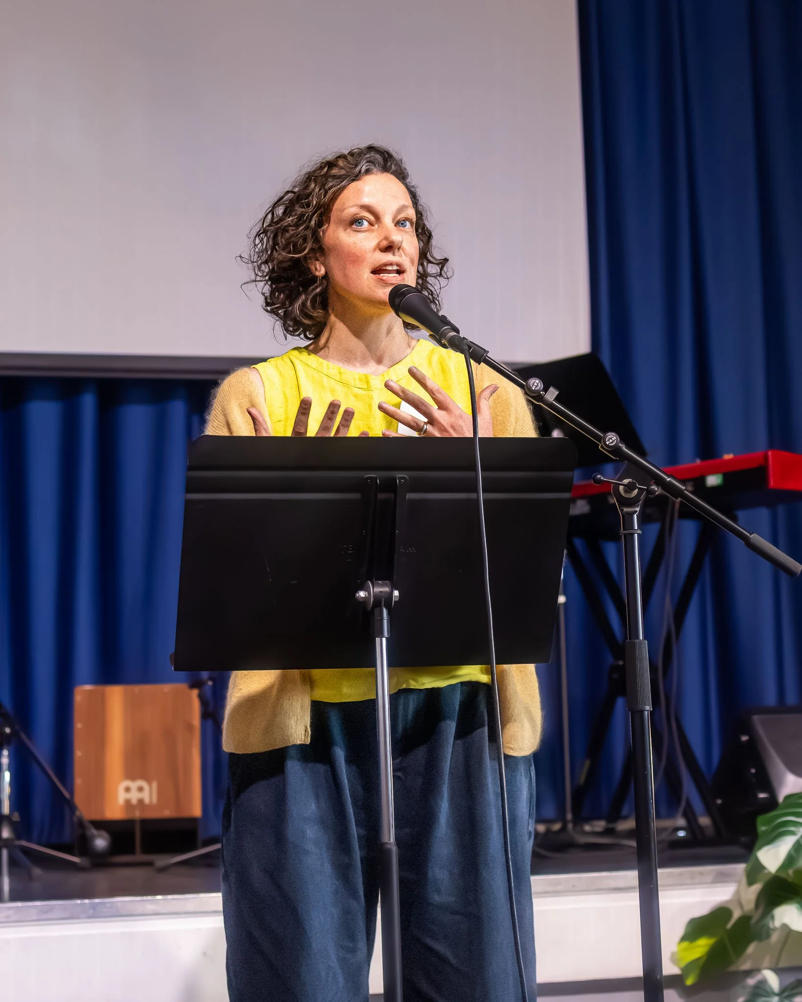 A woman with curly brown hair and blue eyes is speaking at a podium with a microphone in a room with blue curtains and a white wall. She is wearing a yellow top and navy pants.