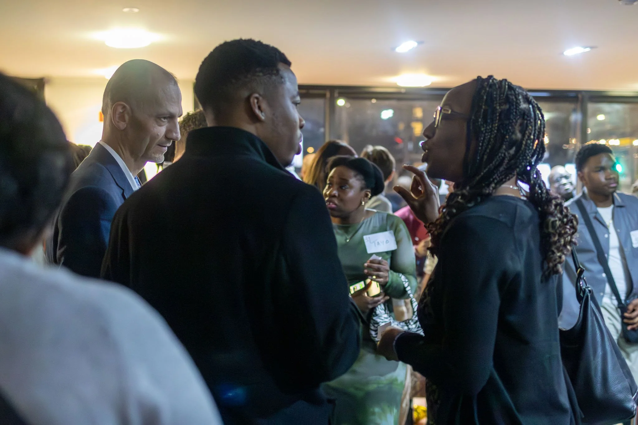 People engaged in conversation at an indoor event, with a woman wearing glasses and a man with a bow tie in the background.