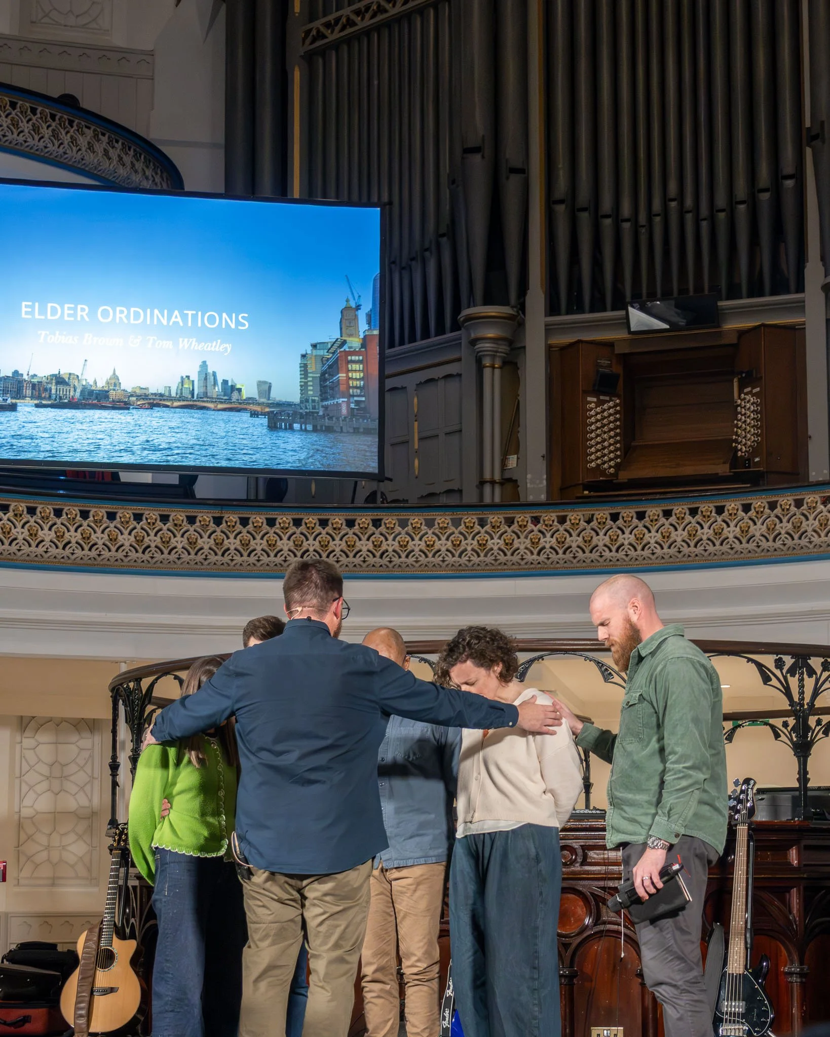 A group of people gathered on a stage during a religious elder ordination ceremony, with a large screen displaying the event title and city skyline in the background.