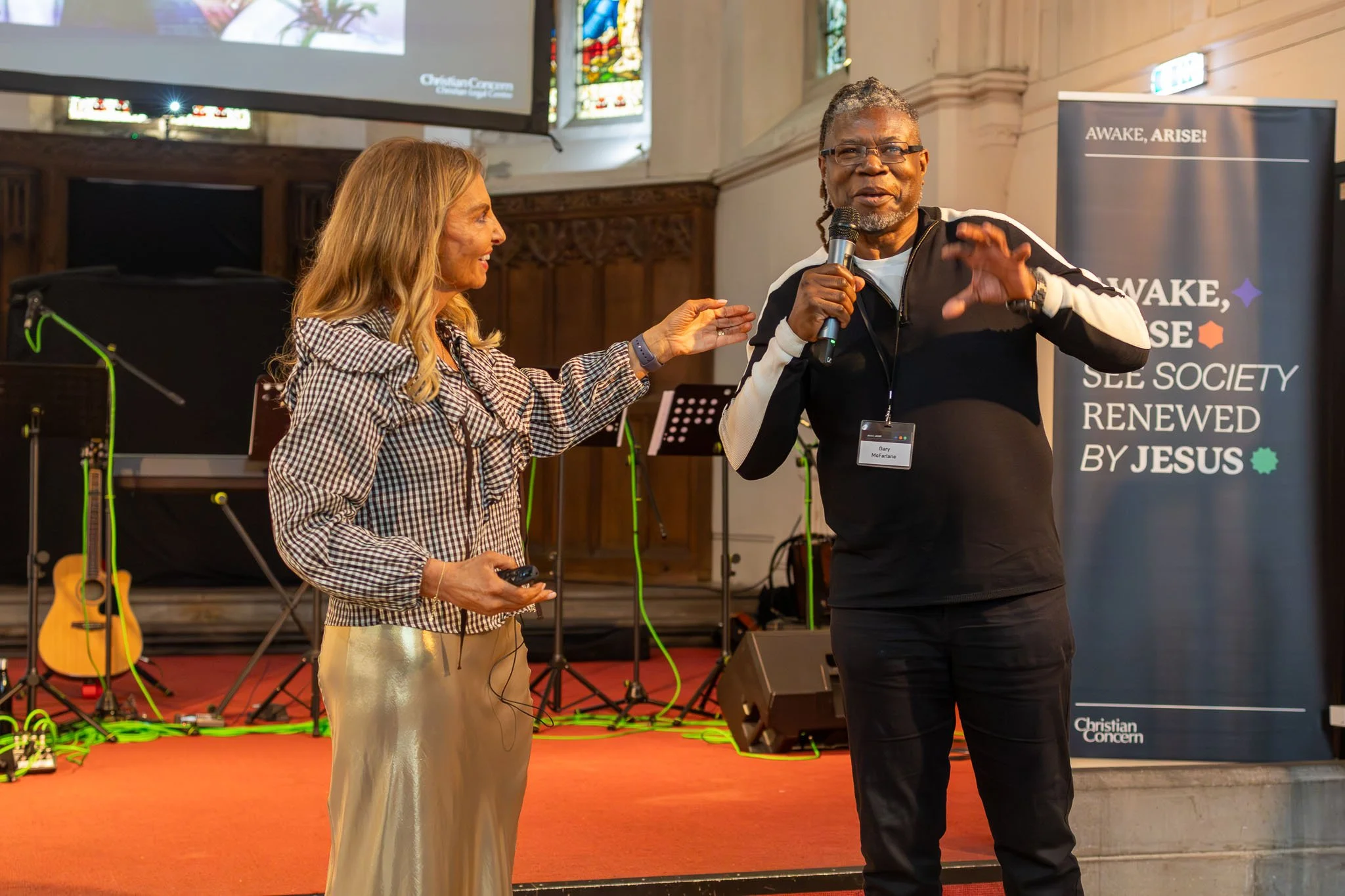 A woman and a man are speaking on stage at an event, with the woman smiling and the man holding a microphone and gesturing with his hand. The woman is wearing a checkered shirt and shiny gold pants, while the man is dressed in a black and white jacke