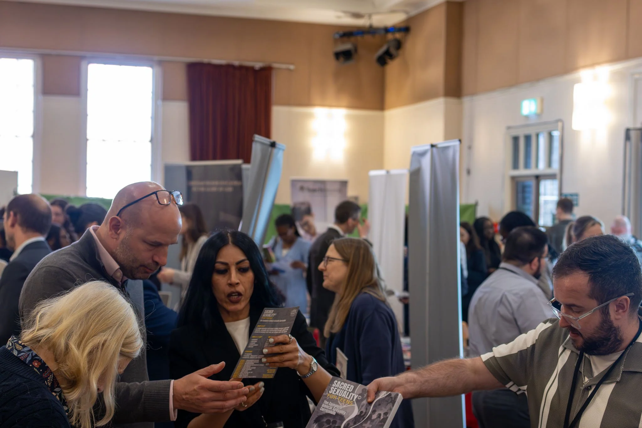 People attending an exhibition at a conference, engaging in conversations and exchanging information in a large room with high windows and wooden walls.