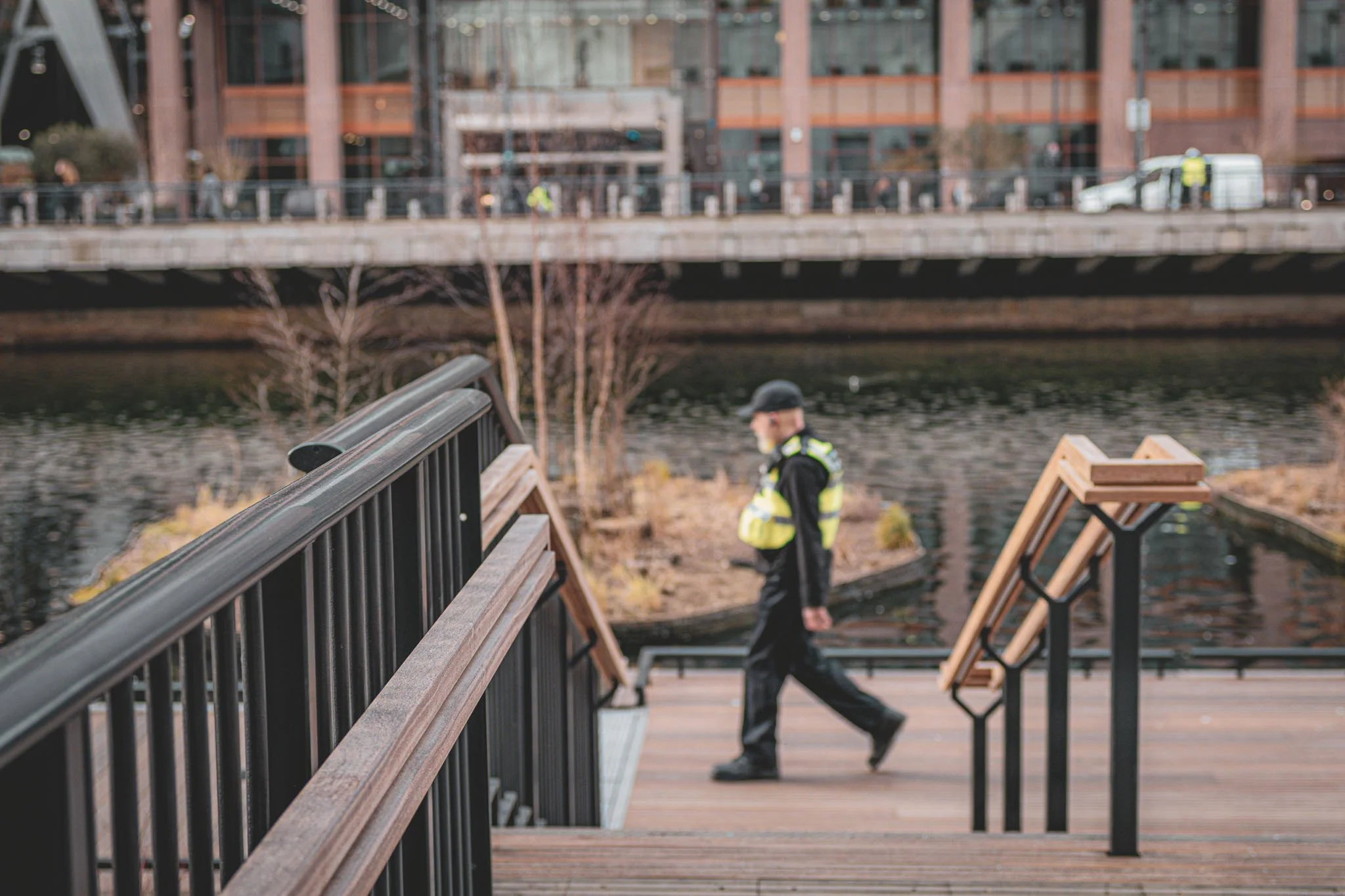 A person in a black jacket and cap walking on a wooden bridge with metal railings, with another person in a yellow vest and cap walking in the background. A building with large windows is visible across the water.