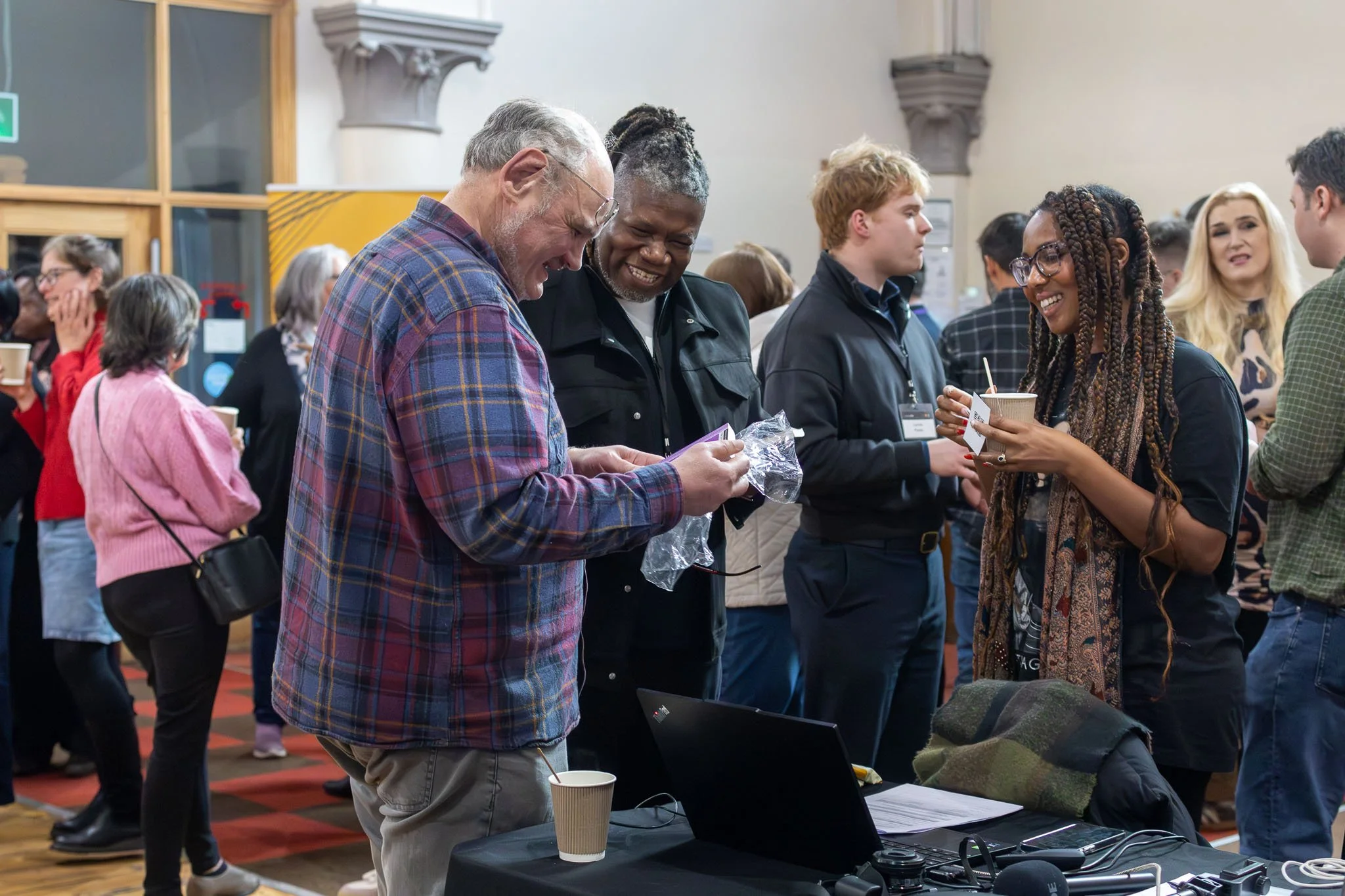 A group of diverse people at a social or networking event, engaging and smiling, with some holding cups and conversing, in an indoor setting.