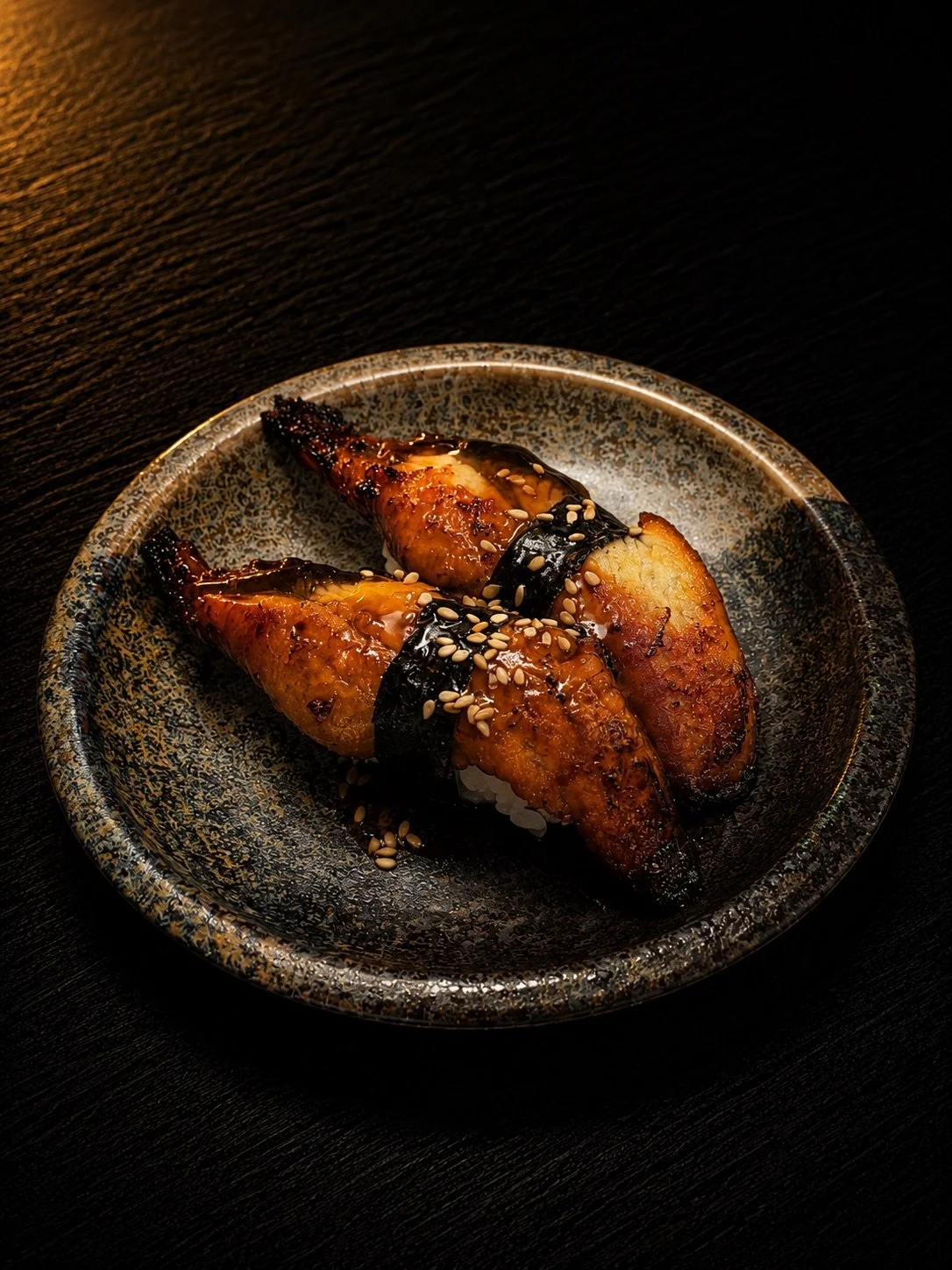 Two pieces of grilled eel sushi topped with sesame seeds, wrapped with seaweed on a ceramic plate