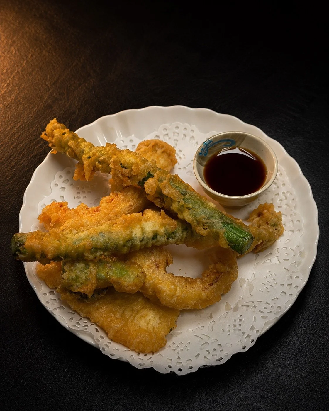 Plate of tempura vegetables, including green beans and sweet potato, with dipping sauce on a white doily-lined plate.